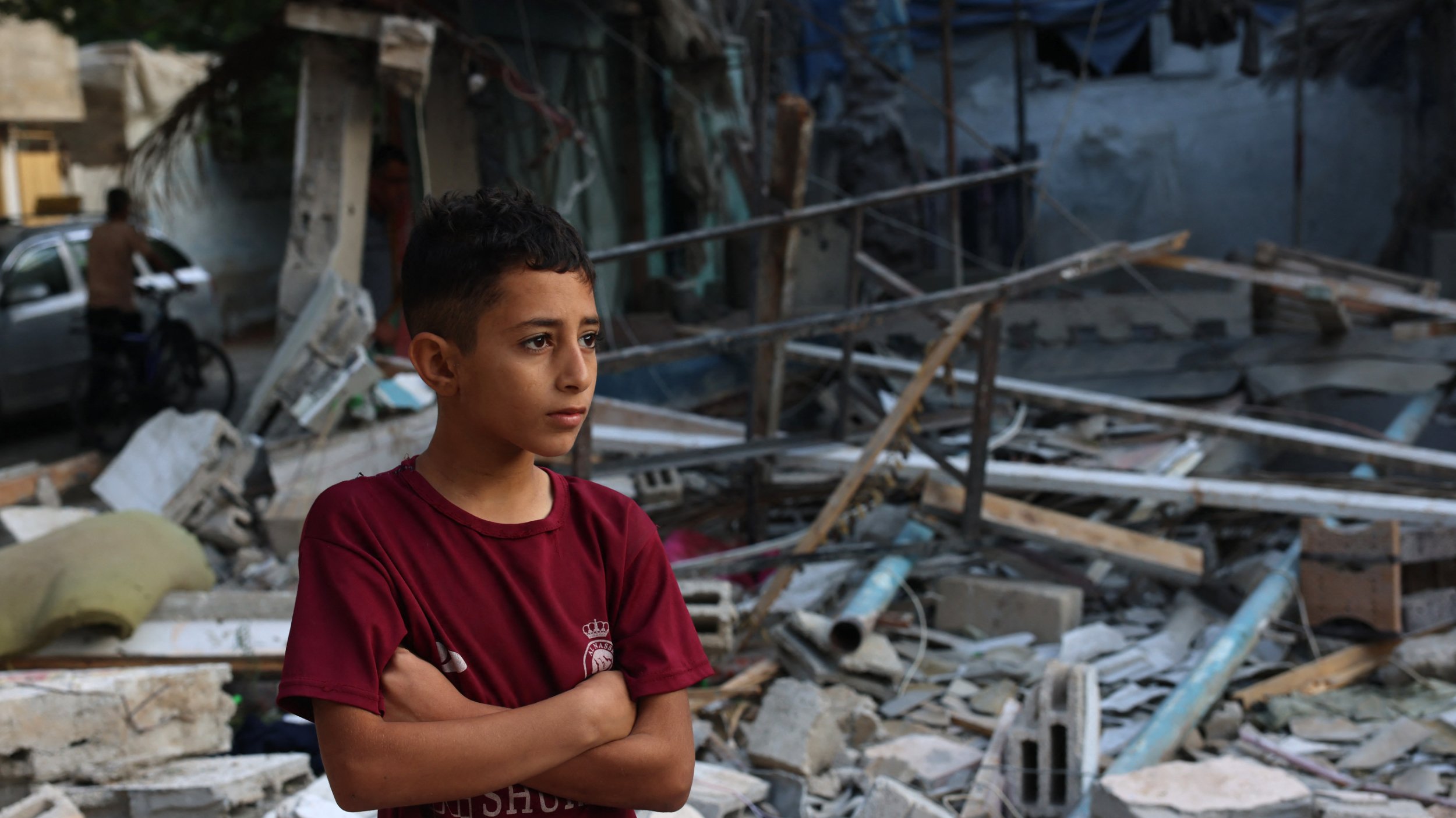 L'image montre un jeune garçon debout, les bras croisés, au milieu des débris d'un bâtiment effondré. Il semble pensif et préoccupé, exprimant une certaine gravité face à la destruction qui l'entoure. Les morceaux de béton et les matériaux épars suggèrent un environnement post-catastrophe, tandis que l'arrière-plan laisse entrevoir d'autres structures endommagées. L'atmosphère générale est sombre et témoigne d'une situation difficile.