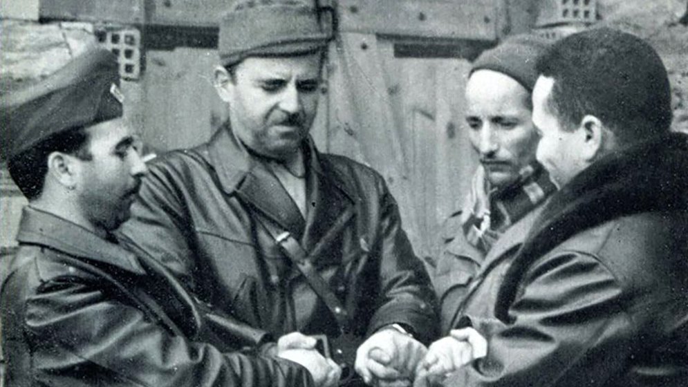 L'image montre quatre hommes serrant les mains les uns des autres dans une posture de camaraderie ou de négociation. Ils portent des tenues militaires ou de combat, et l'environnement semble être extérieur, avec des murs en pierre en arrière-plan. L'expression de leurs visages peut suggérer la détermination ou la concentration, et l'interaction semble exprimer un moment significatif.