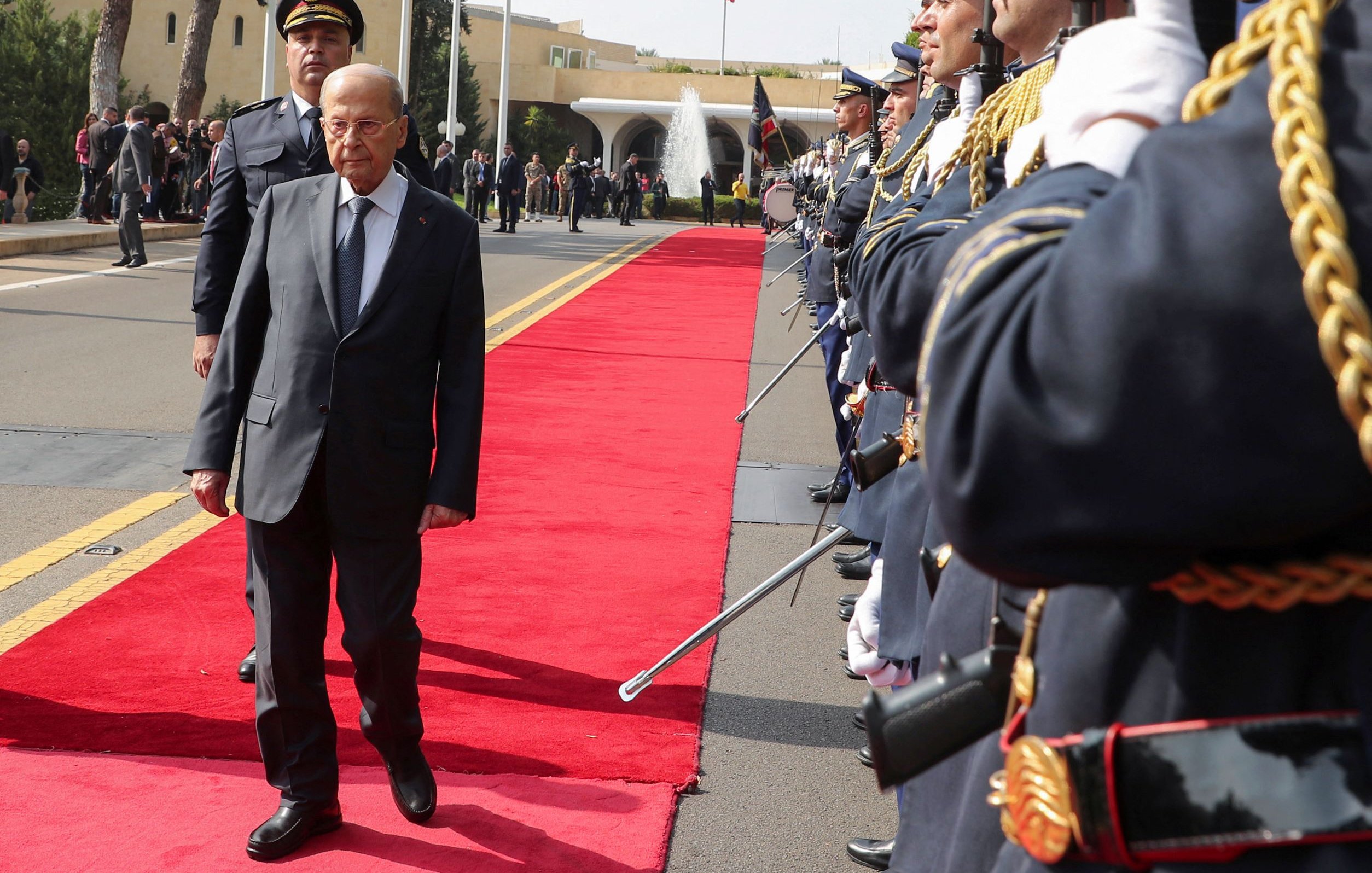 L'image montre une cérémonie officielle. Un homme en costume, probablement une personnalité politique, marche sur un tapis rouge au centre de la scène. De chaque côté, des membres d'une garde d'honneur en uniforme militaire sont alignés, tenant des armes. En arrière-plan, on aperçoit des bâtiments officiels et des drapeaux. L'atmosphère semble solennelle et formelle, suggérant une réception ou un événement diplomatique.