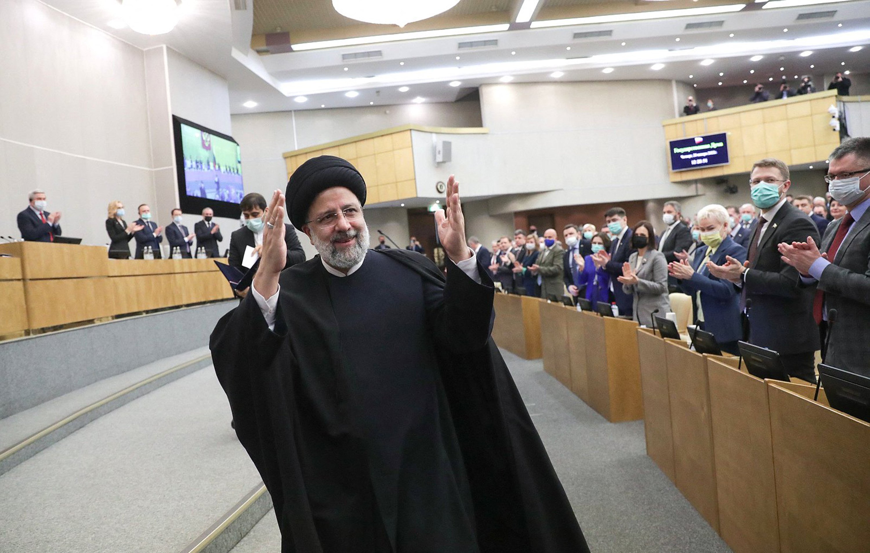 L'image montre un homme en habit traditionnel, portant un turban noir, qui semble être en train de saluer un public. Il se tient au centre d'une salle où plusieurs personnes applaudissent. L'environnement est formel, typique d'un événement politique ou d'une réunion officielle, avec des sièges et des écrans visibles en arrière-plan. Les participants portent également des masques de protection, indiquant une attention à la santé publique.