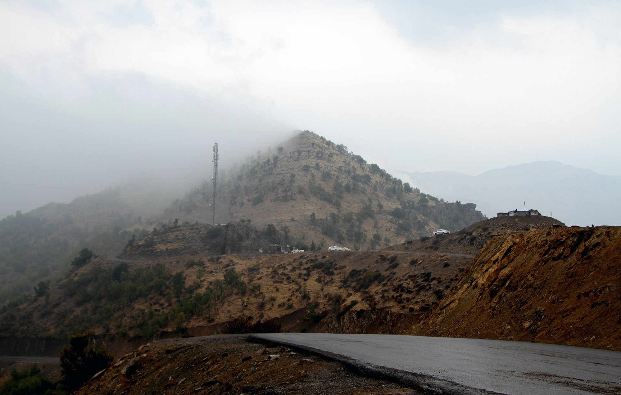 L'image montre un paysage montagneux avec des collines voilées par le brouillard. En arrière-plan, une structure de télécommunication est visible au sommet d'une colline. La route qui serpente est goudronnée, et le terrain environnant présente des nuances de terre brune et de verdure. L'atmosphère semble sombre et mystérieuse, suggérant une journée nuageuse et humide.