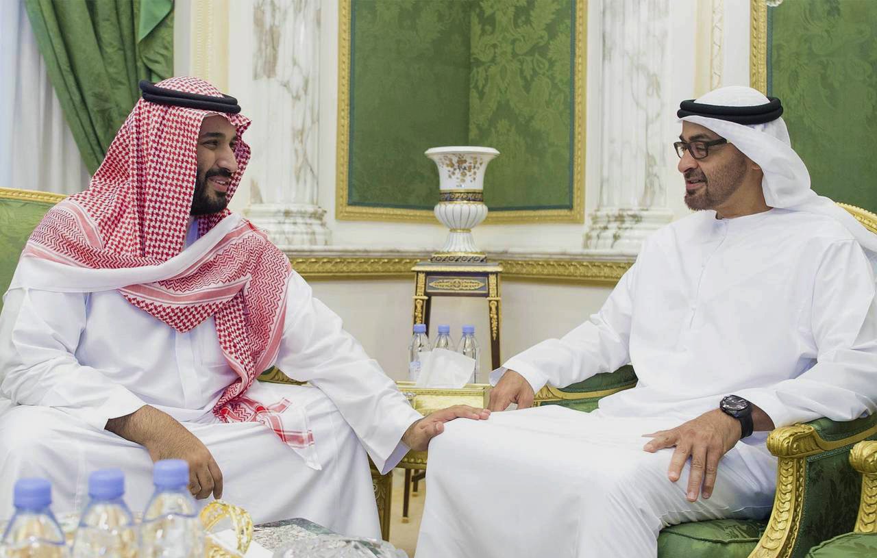 L'image montre deux hommes en vêtements traditionnels. L'un porte une robe blanche avec un manteau rouge et blanc, tandis que l'autre est habillé en blanc avec un couvre-chef noir. Ils sont assis côte à côte sur des fauteuils dans un cadre élégant, décoré de vert et d'or. Les deux hommes semblent engagés dans une conversation amicale. Sur la table devant eux, on aperçoit des bouteilles d'eau.