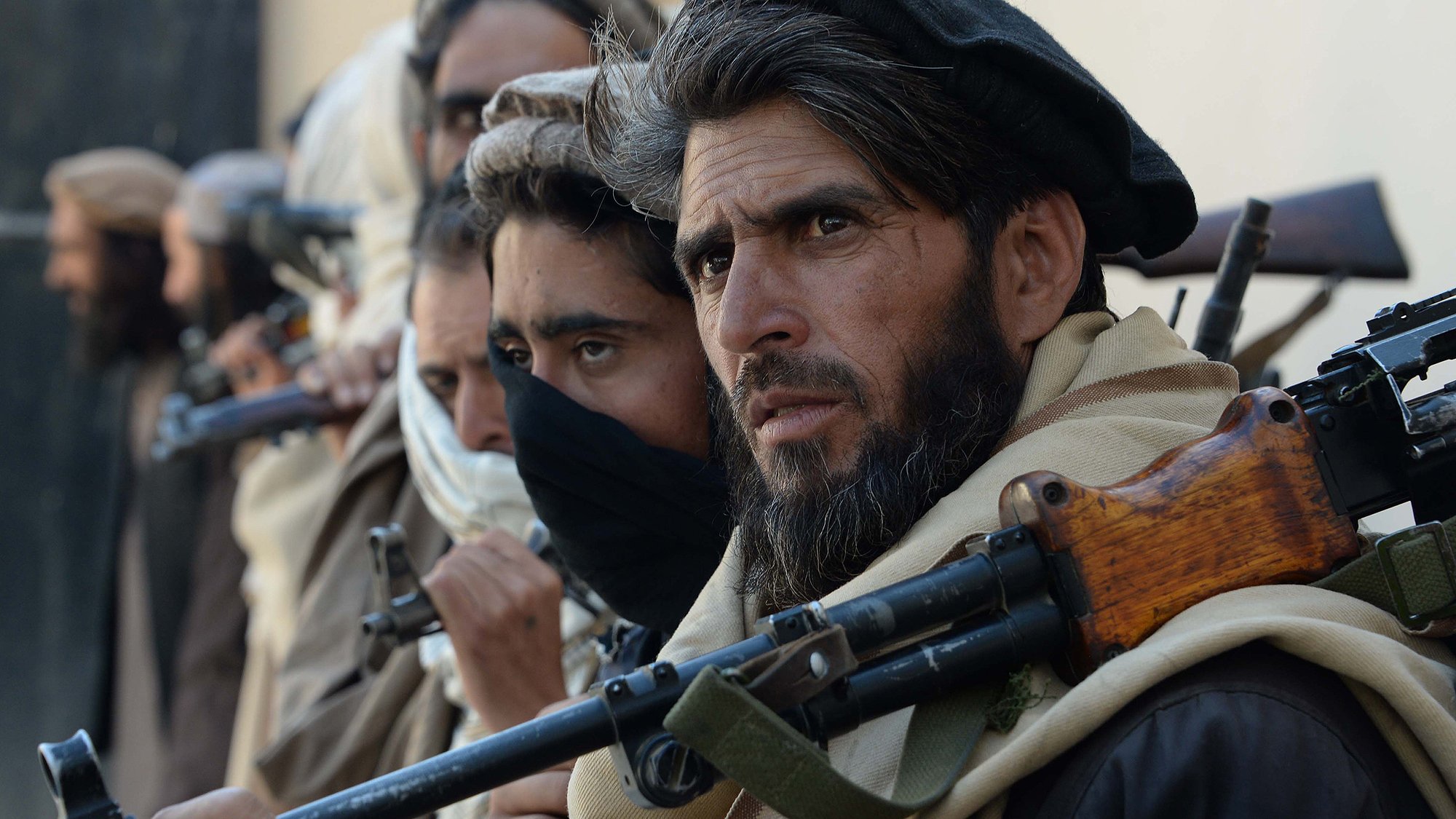 L'image montre un groupe de personnes, apparemment des combattants, qui se tiennent alignés. Ils portent des vêtements traditionnels, certains avec des écharpes ou des masques couvrant leur visage. Chaque individu semble tenir une arme, et leurs expressions sont sérieuses et concentrées. L'environnement semble être urbain ou désertique, avec un fond flou qui suggère une atmosphère de tension.