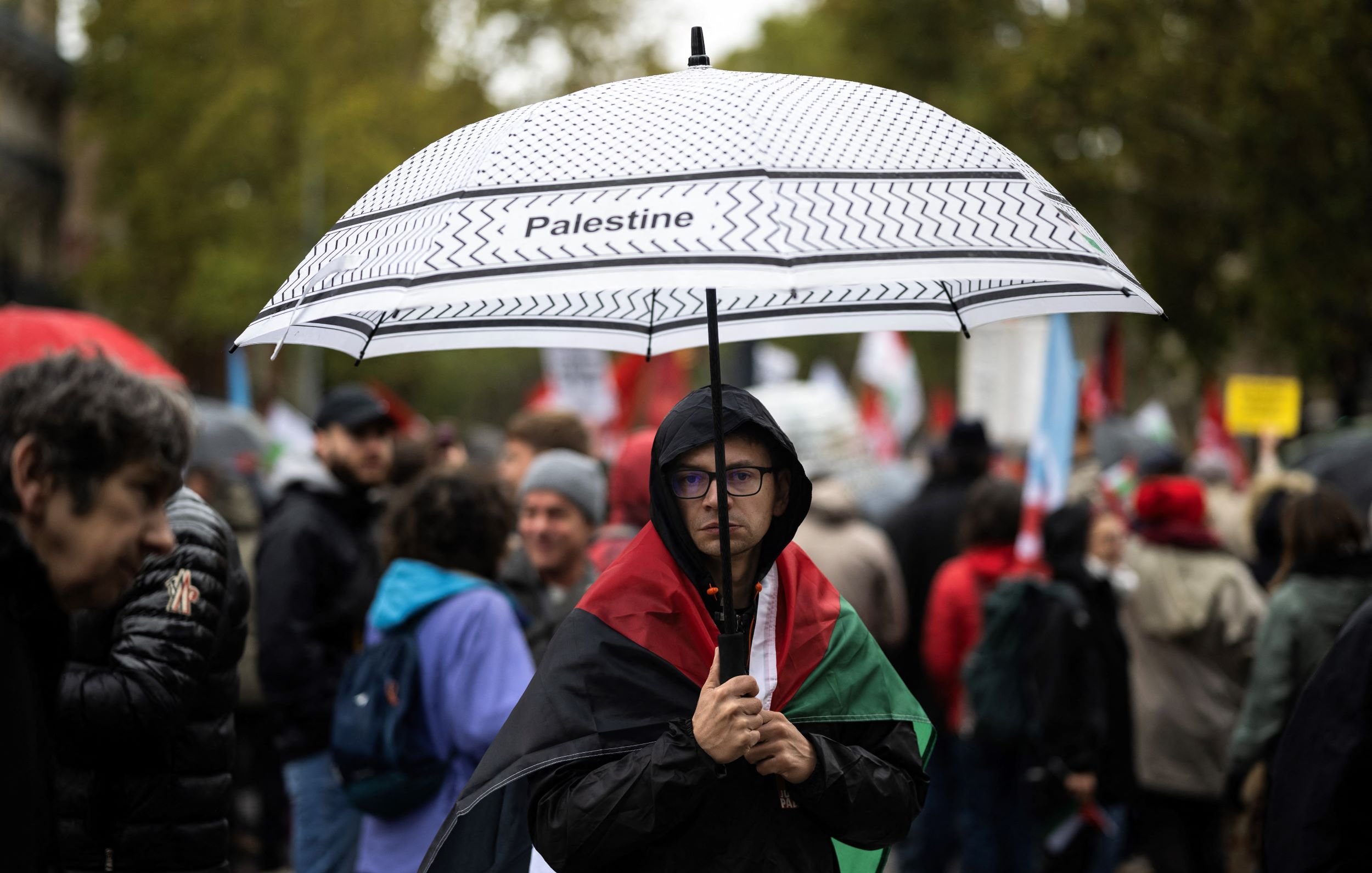 L'image montre une personne portant un parapluie noir et blanc sur lequel est écrit "Palestine". Cette personne est habillée d'un manteau sombre et se tient au milieu d'une foule qui semble participer à une manifestation. En arrière-plan, on peut apercevoir d'autres manifestants, certains portant des drapeaux et des pancartes. L'ambiance semble sérieuse et déterminée, avec des arbres et un ciel nuageux en arrière-plan.