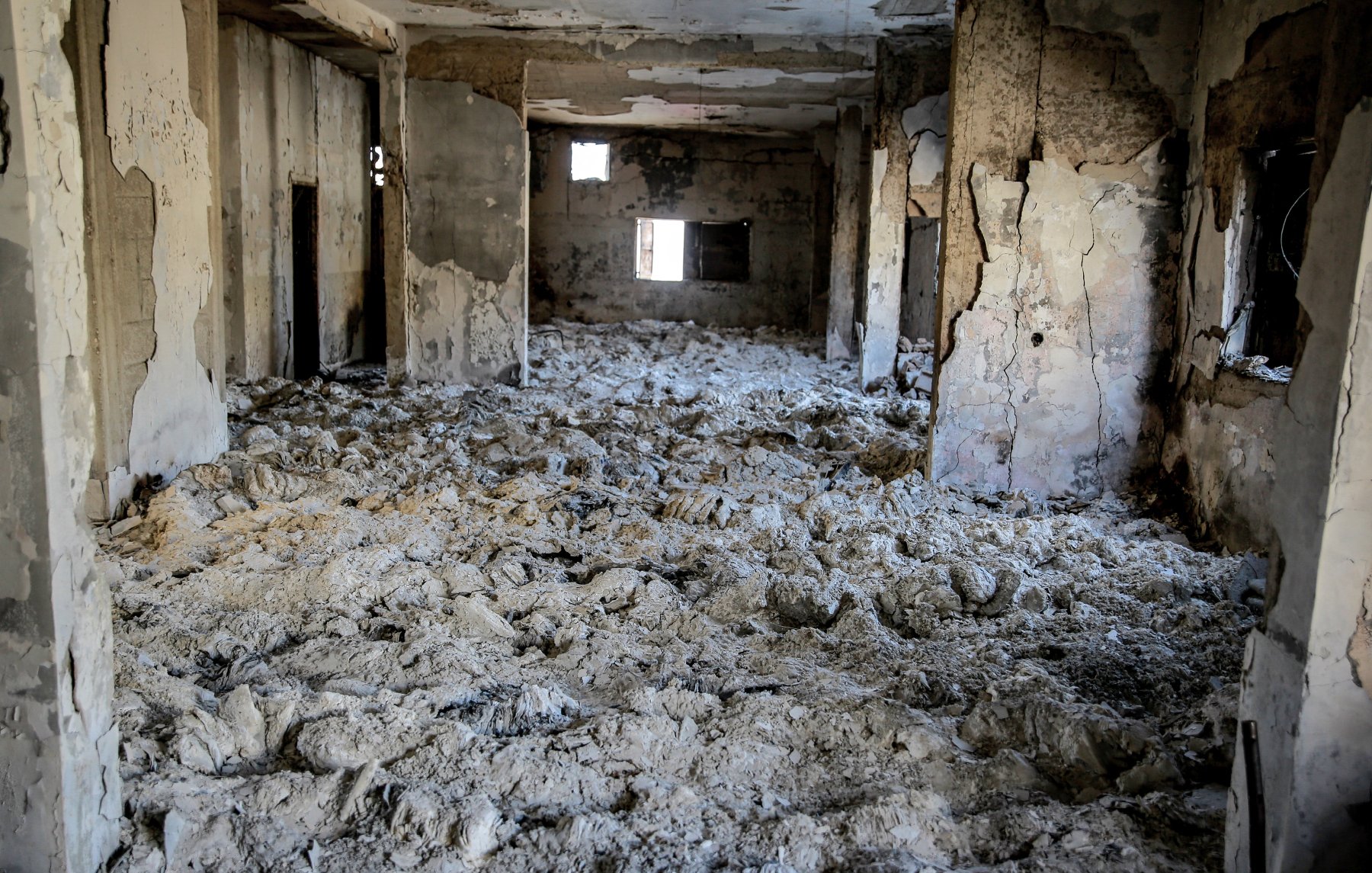L'image montre l'intérieur d'un bâtiment en ruine, où les murs sont endommagés et décrépis. Le sol est recouvert d'un matériau blanc poussiéreux, probablement des débris ou des restes d'anciennes structures. Les fenêtres sont brisées ou manquantes, créant une atmosphère de désolation et d'abandon. La lumière pénètre à travers les ouvertures, mais l'espace parait sombre et sinistre.