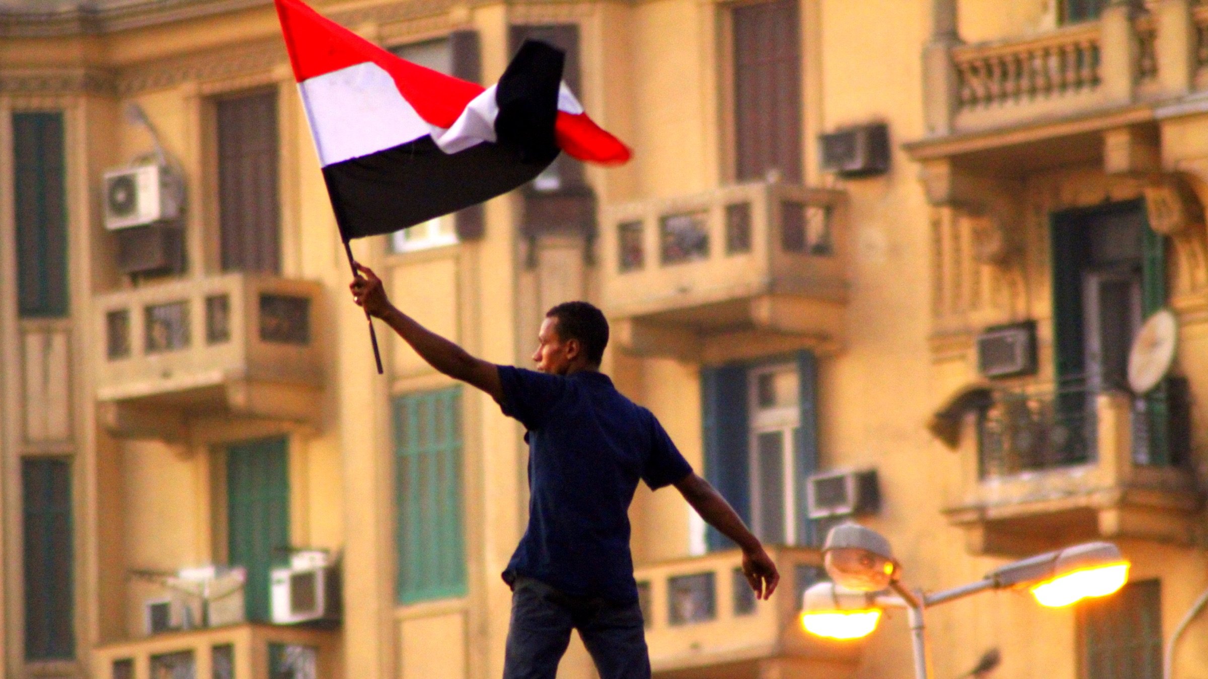 L'image montre un homme brandissant un drapeau noir, blanc et rouge, probablement un drapeau national. Il se tient debout sur une structure, manifestant une posture de célébration ou de protestation. En arrière-plan, on peut apercevoir des bâtiments avec des balcons et des climatiseurs, ce qui suggère un environnement urbain. L'atmosphère semble chargée d'une forte émotion, possiblement liée à un événement politique ou social.