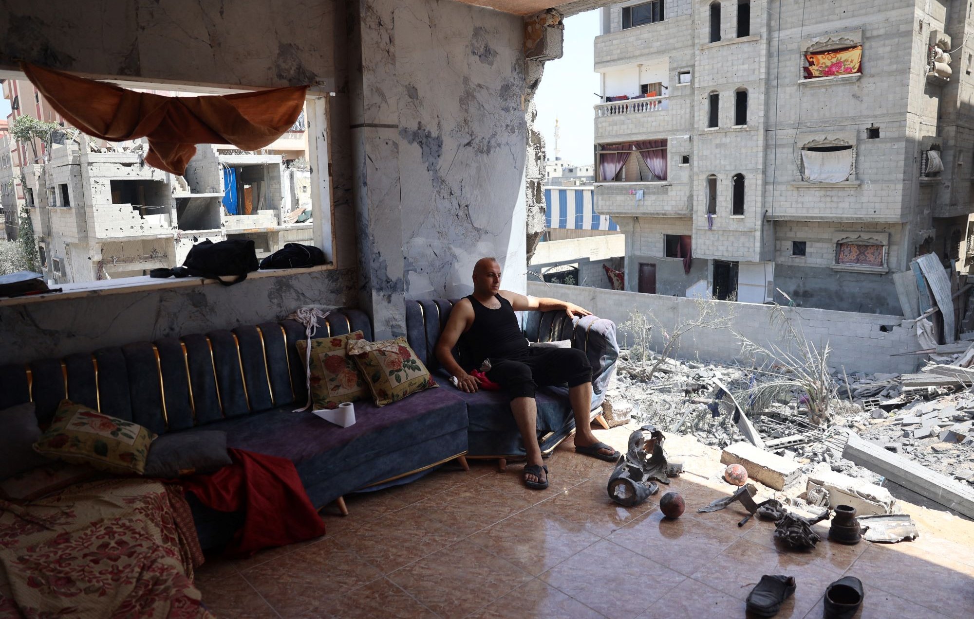 L'image montre un homme assis sur un canapé dans une pièce endommagée. Il semble se reposer, vêtu d'un débardeur noir et d'un pantalon court et noir. La pièce est en mauvais état, avec des murs endommagés et des débris au sol. À l'extérieur de la pièce, on aperçoit des bâtiments gravement endommagés, suggérant qu'il s'agit d'une zone touchée par un conflit ou une catastrophe. Le mobilier à l'intérieur, comme le canapé et certains coussins, semble également en désordre. L'atmosphère générale est celle de la dévastation et de la récupération après un événement destructeur.