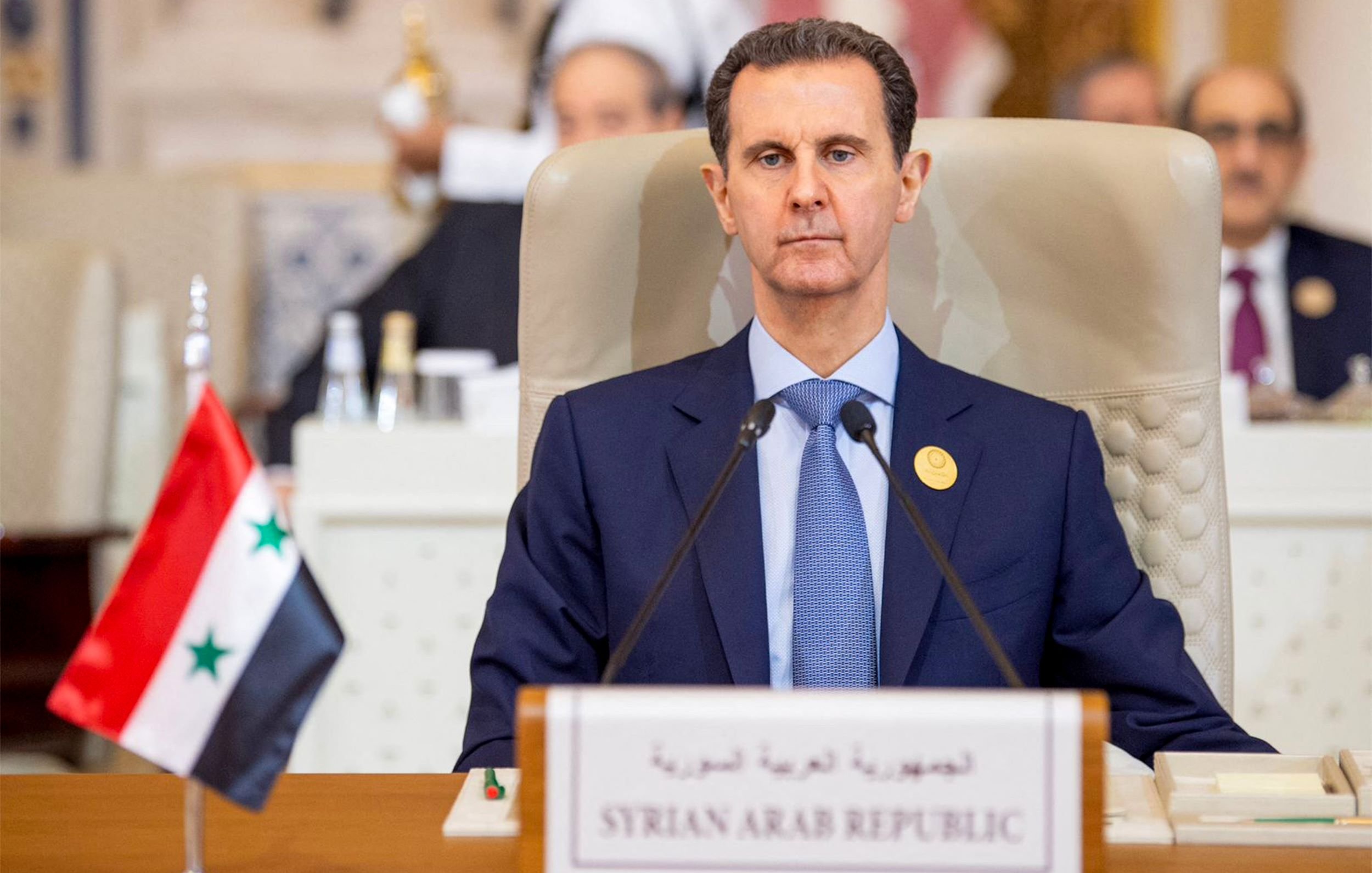 L'image montre un homme assis derrière un bureau lors d'une réunion officielle. Il est habillé en costume sombre et semble sérieux. À côté de lui, il y a un drapeau de la Syrie et une plaque indiquant "République arabe syrienne". En arrière-plan, on peut apercevoir d'autres personnes et une disposition formelle typique d'une conférence ou d'un sommet.