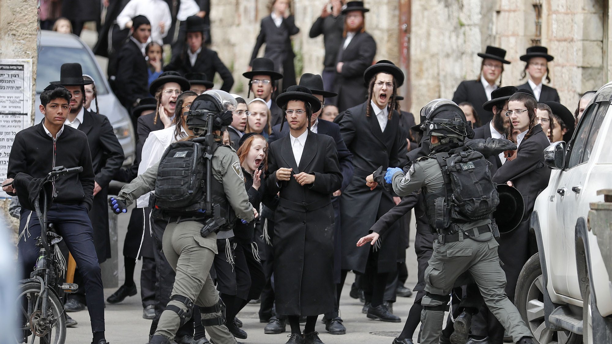 L'image montre une scène animée dans une rue, où un groupe de personnes, principalement habillées en noir, semble manifester ou réagir à une situation tendue. On peut voir des hommes en habits traditionnels, portant des chapeaux noirs, ainsi que des adolescents sur des bicycles. Au centre, des membres des forces de l'ordre, en tenue tactique, interagissent avec la foule. Les expressions faciales des personnes dans la foule varient entre la surprise, l'inquiétude et l'agitation, créant une atmosphère de tensions. Les bâtiments en arrière-plan évoquent un contexte urbain, probablement dans une région où se concentrent des communautés religieuses.