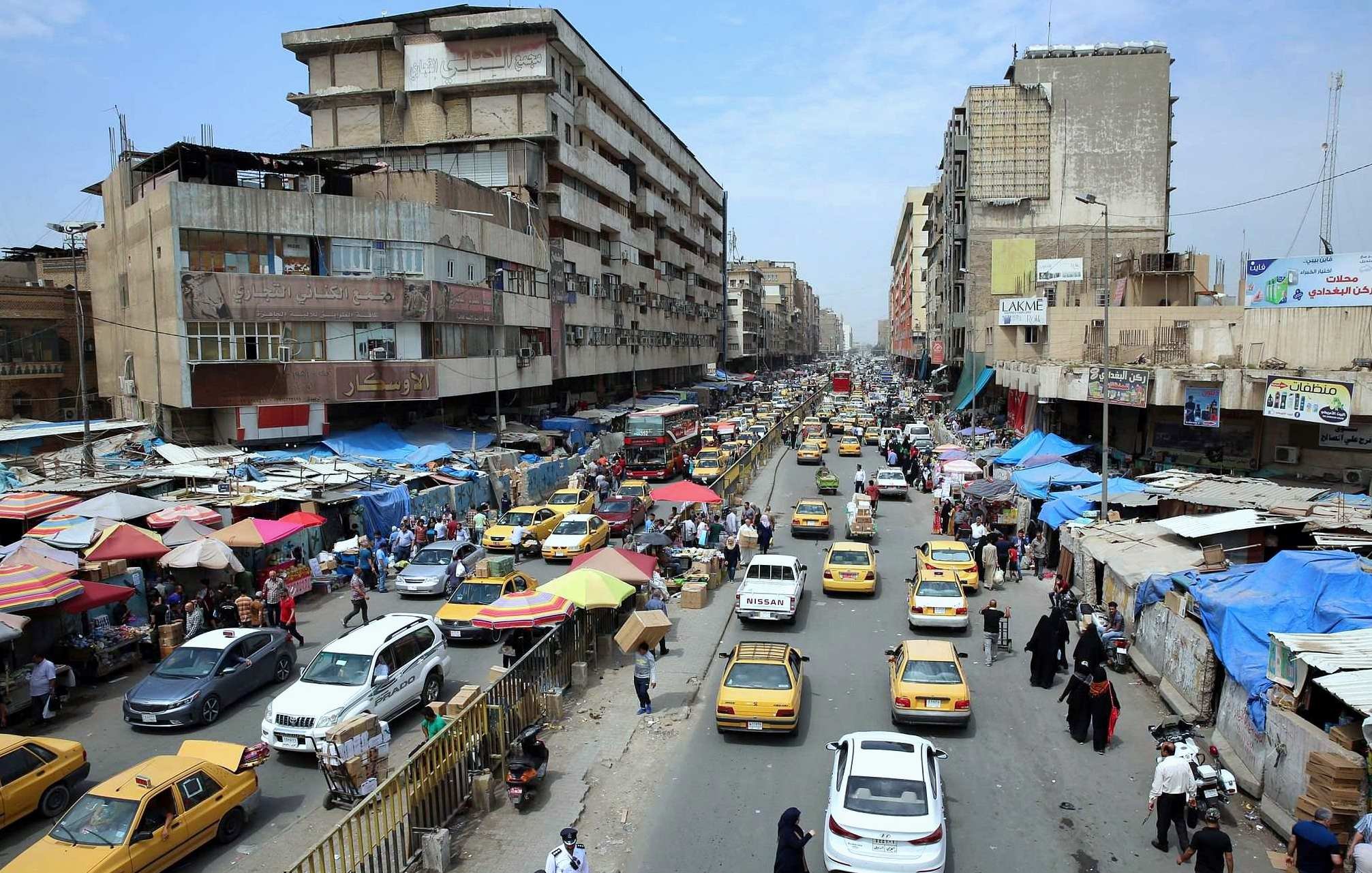 L'image présente une scène urbaine animée, probablement dans une grande ville. On peut voir une rue bordée de bâtiments qui semblent vieux et usés. La circulation est dense, avec de nombreux taxis jaunes qui circulent dans les deux sens. Des échoppes et des marchés de rue sont visibles, avec des auvents colorés, indiquant une ambiance de commerce local. Les gens marchent le long des trottoirs, et l'ensemble de la scène donne une impression de mouvement et de vie citadine. Le ciel est partiellement nuageux, ce qui suggère un climat chaud.