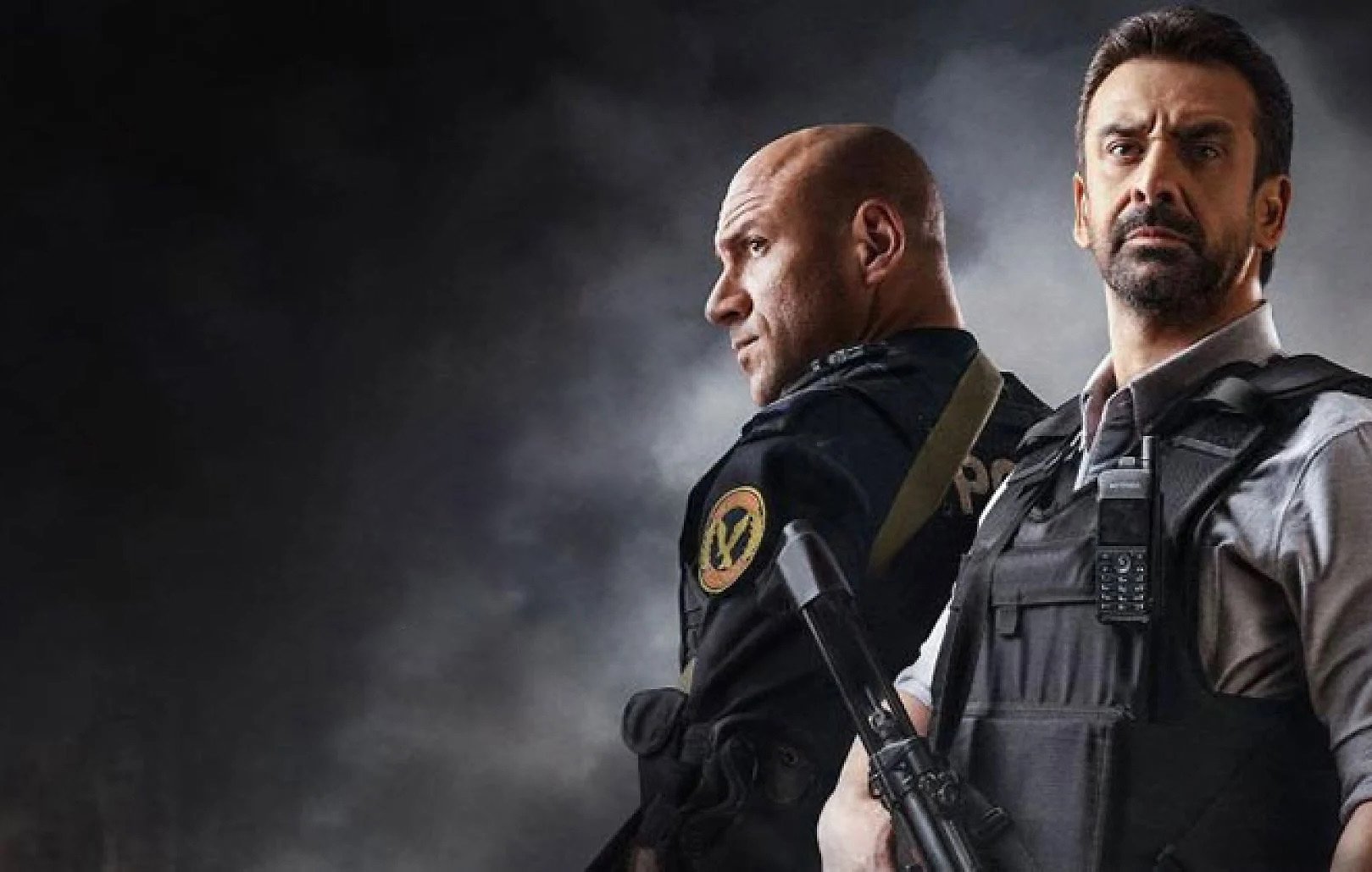 L'image montre deux hommes en tenue tactique, se tenant dos à dos dans une ambiance sombre et intense. L'un d'eux porte un uniforme de police ou militaire, tandis que l'autre est habillé d'une chemise et d'un gilet pare-balles. Ils semblent prêts pour une situation d'urgence, chacun tenant une arme à feu. Le fond est flou et dramatique, ajoutant à la tension de la scène.