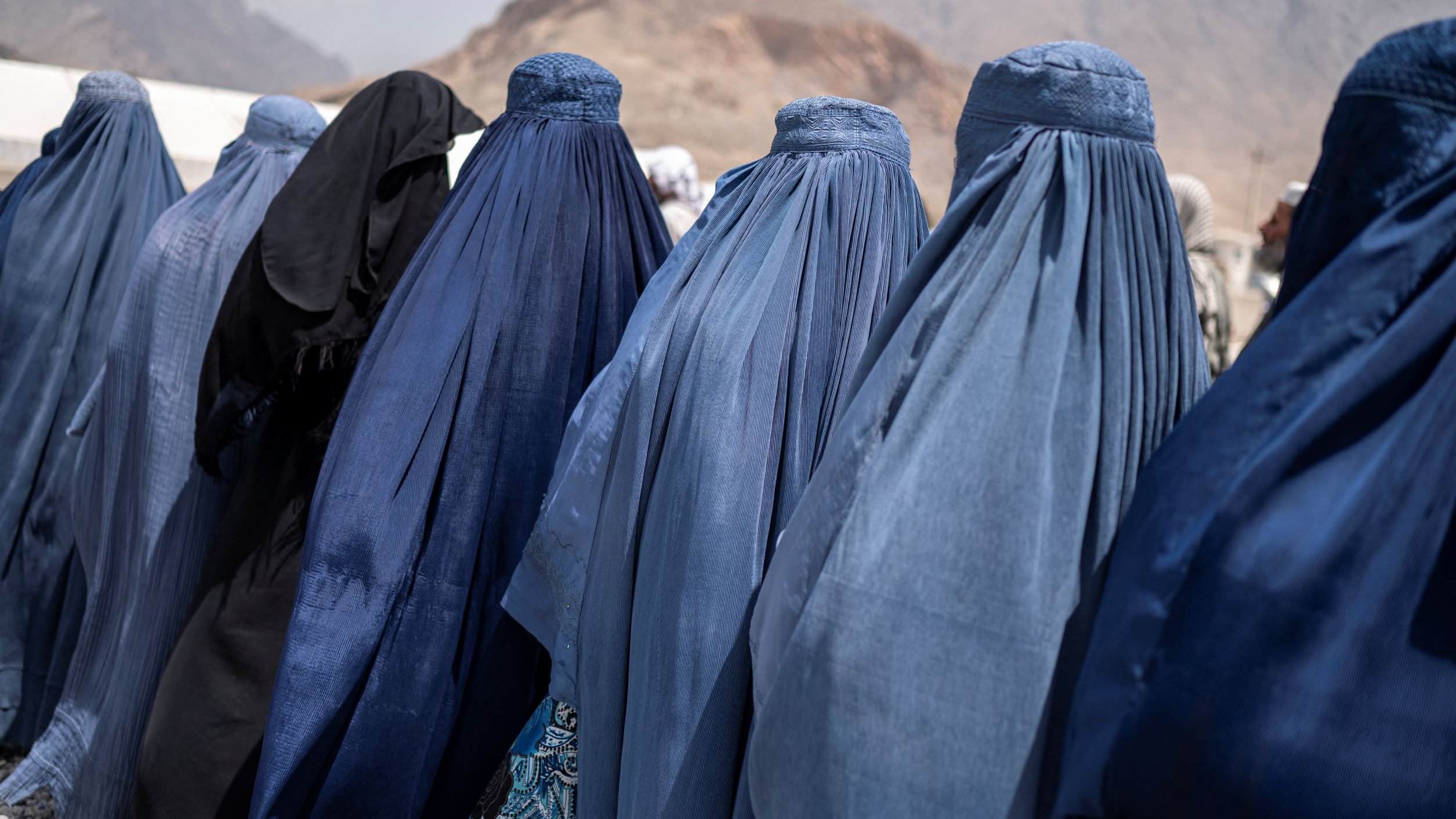 L'image montre un groupe de femmes de dos, vêtues de burqas de différentes nuances de bleu, se tenant en ligne. On peut également apercevoir une femme portant des vêtements noirs. En arrière-plan, on distingue des montagnes, et le ciel semble nuageux. L'ambiance générale semble être celle d'un rassemblement ou d'une rencontre.