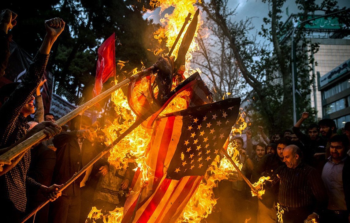 L'image montre une scène de protestation où des gens brûlent un drapeau américain. Les participants, visiblement passionnés, lèvent les bras et brandissent des bâtons. L'atmosphère est intense, avec des flammes qui s'élèvent autour du drapeau, tandis que des arbres et des bâtiments en arrière-plan ajoutent à la dynamique de la situation. Cette action symbolise souvent une forte opposition ou un message politique.