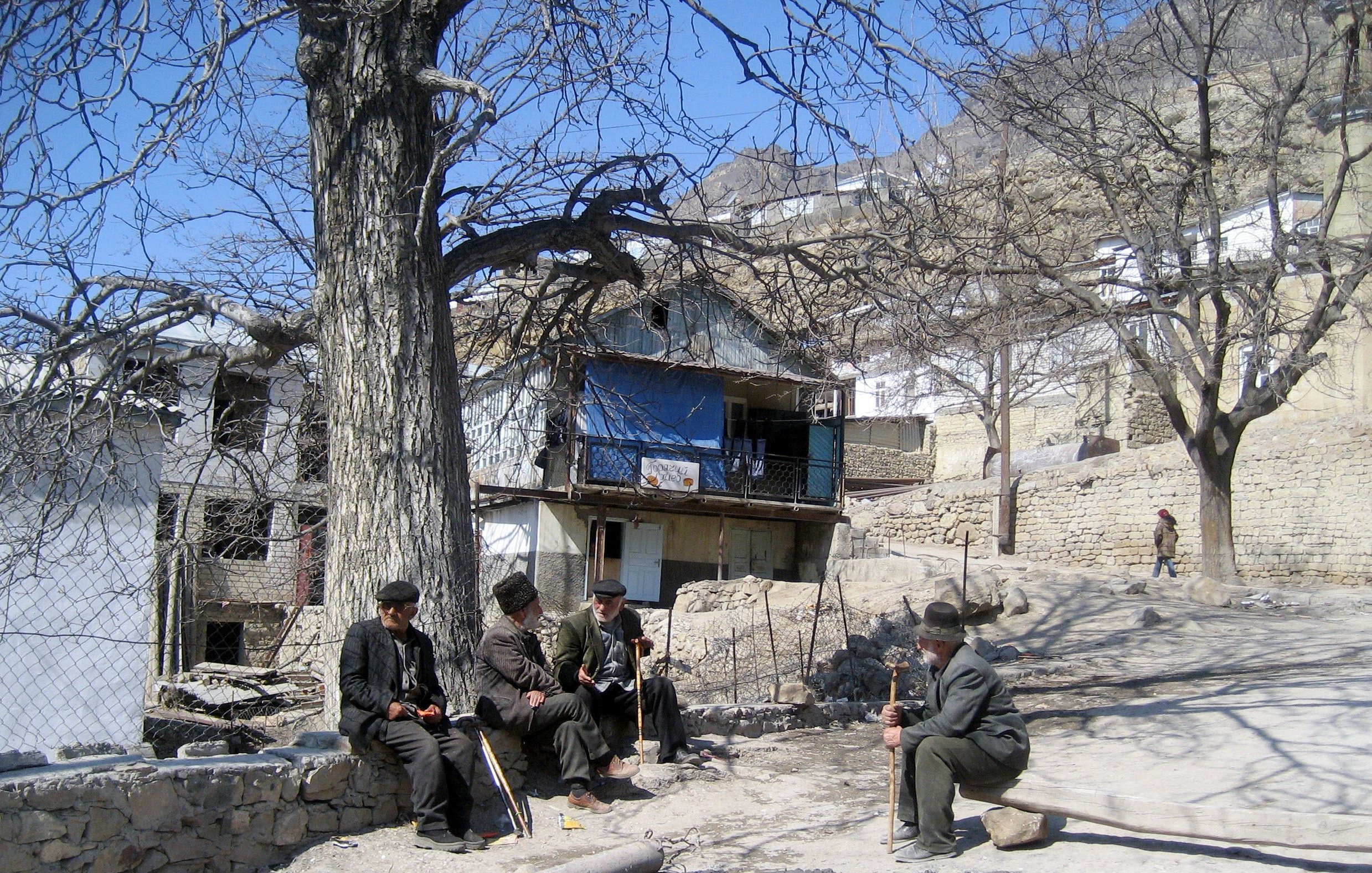 L'image montre un paysage rural avec des maisons en pierre, certaines d'entre elles ayant des balcons. Au premier plan, quatre hommes assis discutent ensemble, habillés de manière traditionnelle. Ils sont entourés d'arbres dénudés, ce qui indique probablement le début du printemps ou l'hiver. L'ambiance semble calme et tranquille, avec un chemin en terre qui serpente entre les maisons.