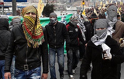 L'image montre une manifestation de foule, où de nombreuses personnes marchent ensemble. Beaucoup d'entre elles portent des masques ou des foulards qui couvrent leur visage, ce qui donne un aspect uniformisé à ce groupe. On constate une ambiance de protestation, probablement liée à une revendication sociale ou politique. En arrière-plan, on aperçoit des drapeaux et des banderoles, indiquant des messages forts. Les participants semblent déterminés, et l'atmosphère semble chargée d'énergie et de tensions.