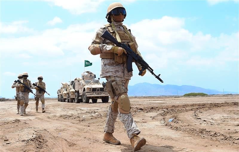 L'image montre des soldats en tenue militaire marchant sur un terrain désertique. Ils portent des casques et des gilets pare-balles, et certains utilisent des armes. En arrière-plan, on peut voir des véhicules militaires. Le ciel est clair, et le paysage est aride. La scène évoque une ambiance de formation ou d'exercice militaire.