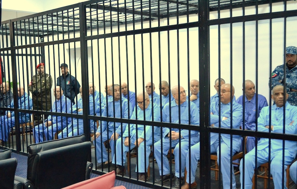 L'image montre un groupe d'individus assis derrière des barreaux. Ils portent tous des vêtements similaires, de couleur bleue. L'atmosphère semble sérieuse et formelle, peut-être en lien avec une audience ou un procès. Plusieurs personnes en uniforme sont présentes, suggérant un contexte de sécurité ou d'autorité. Les individus semblent concentrés ou préoccupés.