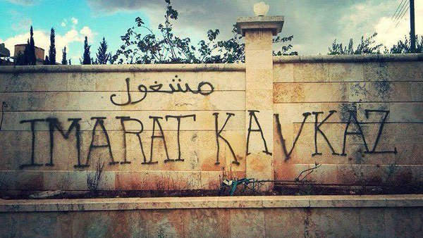 L'image montre un mur en pierres, probablement d'un bâtiment ou d'une clôture. Sur ce mur, il y a des graffitis qui sont en arabe et en lettres latines. Le mot en arabe semble se traduire par "travail" ou "emploi", tandis que "IMARAT KAVKAZ" est écrit en grandes lettres au centre. L'environnement autour du mur est nature, avec quelques arbres et un ciel nuageux, ce qui donne une ambiance un peu mélancolique ou chargée.