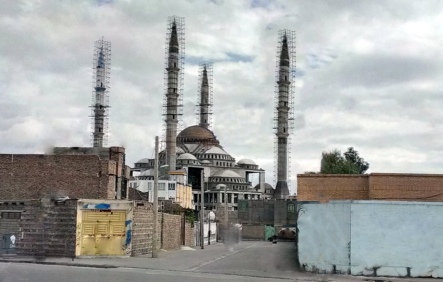 L'image montre un grand bâtiment au centre, semblable à une mosquée, avec plusieurs minarets élancés qui s'élèvent vers le ciel. Le bâtiment a un dôme proéminent et semble être en construction ou en rénovation, avec des échafaudages visibles. En arrière-plan, le ciel est nuageux, ce qui donne à la scène une atmosphère un peu sombre. À l'avant, on peut apercevoir des structures et des murs en briques, ainsi qu'une route. L'ensemble de la composition évoque un mélange d'architecture traditionnelle et moderne.
