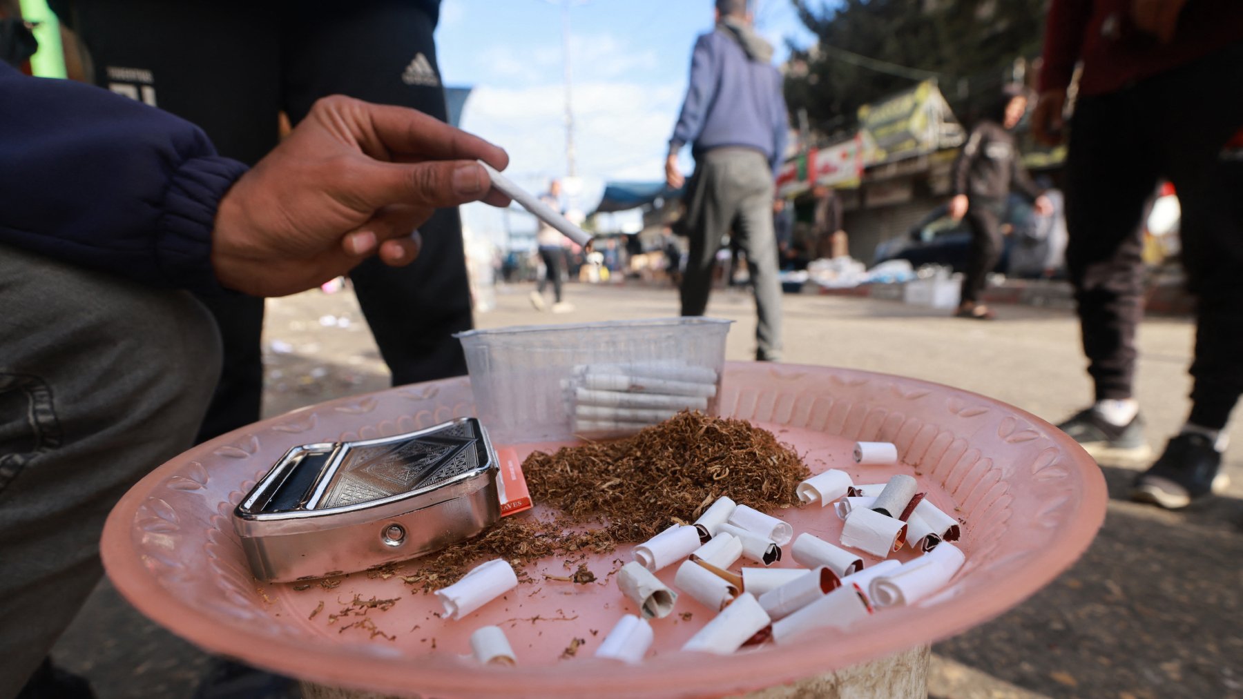 L'image montre une scène de rue avec une personne qui tient une cigarette à la main. Sur un plateau, on peut voir du tabac en vrac, des cigarettes déjà consommées et un petit récipient. En arrière-plan, on aperçoit des passants et des affiches sur des murs. L'ambiance semble animée et urbaine.