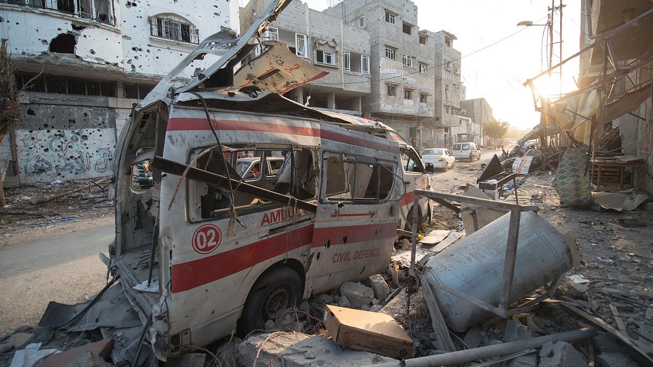 L'image montre une scène de destruction urbaine, probablement à la suite d'un conflit. On y voit une ambulance endommagée, avec sa structure déformée et son revêtement extérieur marqué par des débris. Les bâtiments environnants sont en ruine, avec des fenêtres brisées et des murs partiellement effondrés. Le sol est jonché de débris, et l'éclairage indique que c'est soit tôt le matin, soit en fin d'après-midi. L'atmosphère générale est sombre et témoigne de l'impact des violences sur la ville.