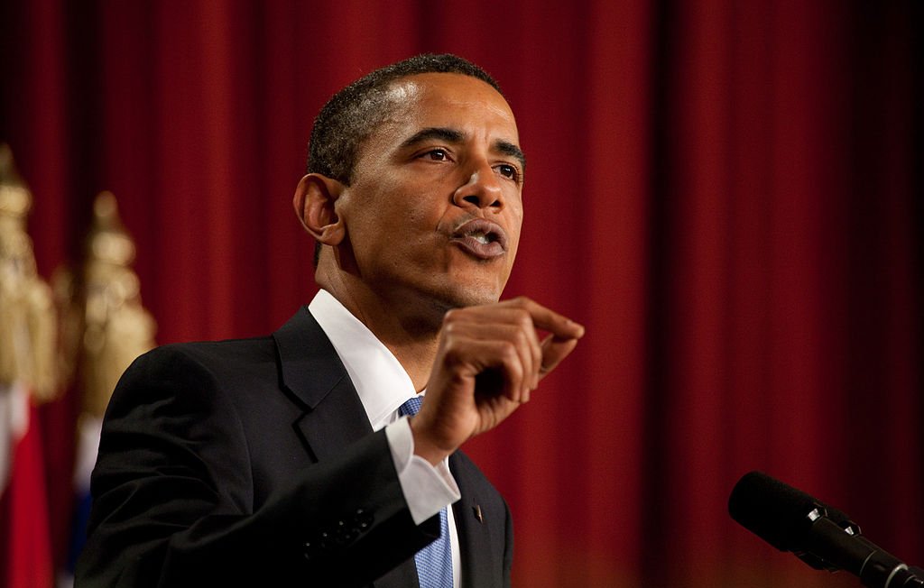 L'image montre un homme en costume noir avec une chemise blanche et une cravate bleue. Il est en train de parler avec passion, levant un doigt pour souligner un point important. En arrière-plan, on peut voir un drapeau ou un décor formel. L'atmosphère semble très sérieuse et engageante, suggérant qu'il est en train de prononcer un discours ou de faire une déclaration importante.