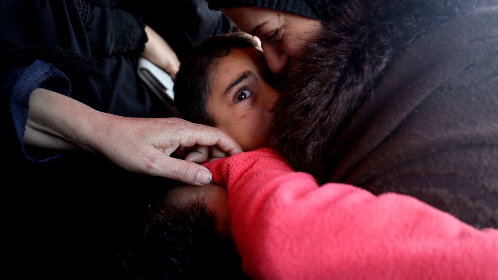 L'image montre un moment touchant entre un enfant et un adulte. L'adulte semble embrasser l'enfant, qui a l'air attentionné et gaze directement vers l'objectif. On peut aussi voir quelques mains, une d'elles touchant l'épaule de l'enfant. L'ambiance est empreinte d'émotion et de tendresse. Les vêtements des figures suggèrent un environnement austère.