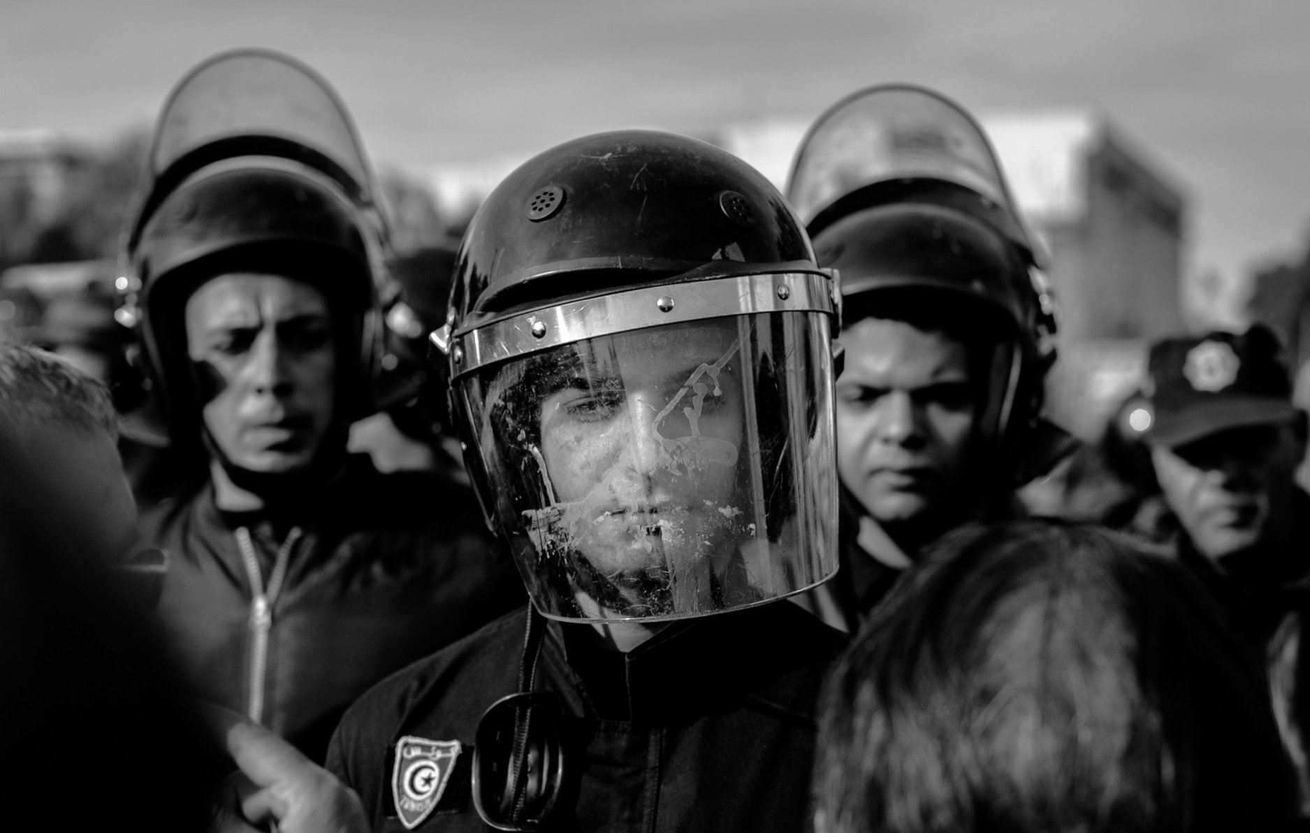L'image montre un groupe de personnes, probablement des agents des forces de l'ordre, entourés par une foule. L'un des agents est au premier plan, portant un casque et un bouclier facial, ce qui indique qu'il est en position de maintien de l'ordre. Les visages des autres personnes en arrière-plan sont également visibles, exprimant une certaine tension. L'image est en noir et blanc, ce qui accentue le sérieux de la situation. L'atmosphère semble chargée, avec un sentiment de confrontation possible.