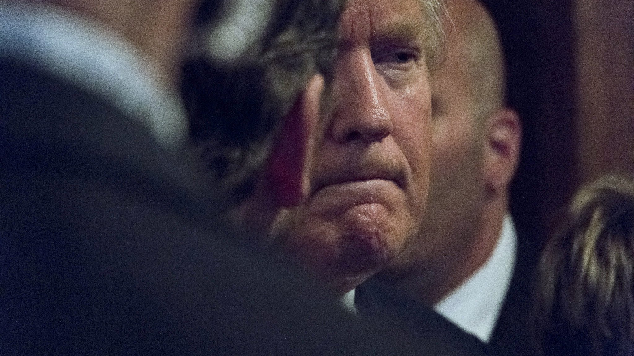 L'image montre un homme avec des cheveux blonds, affichant une expression pensive ou sérieuse. Il est partiellement obscurci par des personnes en arrière-plan, ce qui crée une atmosphère de concentration ou d'intensité. On peut également remarquer des costumes sombres, suggérant un cadre formel.