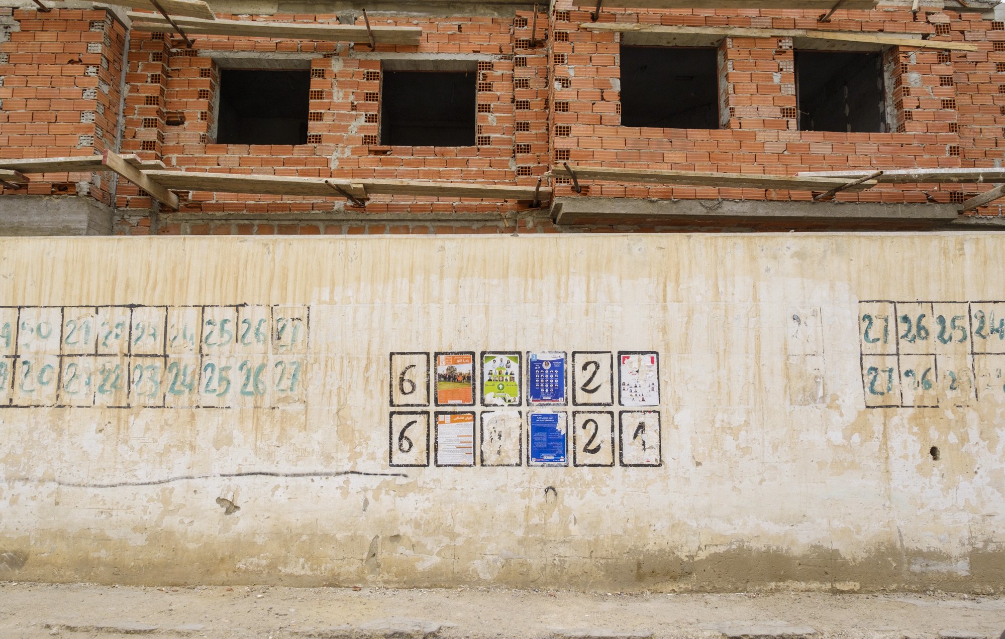 L'image montre un mur en béton, partiellement recouvert de peinture, qui semble faire partie d'un chantier de construction. Sur ce mur, il y a plusieurs cases carrées, probablement marquées pour indiquer des numéros ou des étapes de progression. À la centre, quelques affiches et documents sont accrochés, fournissant des informations peut-être liées au chantier. En arrière-plan, on aperçoit un bâtiment en construction avec des briques exposées et des fenêtres incomplètes, ce qui suggère un contexte de travaux en cours.