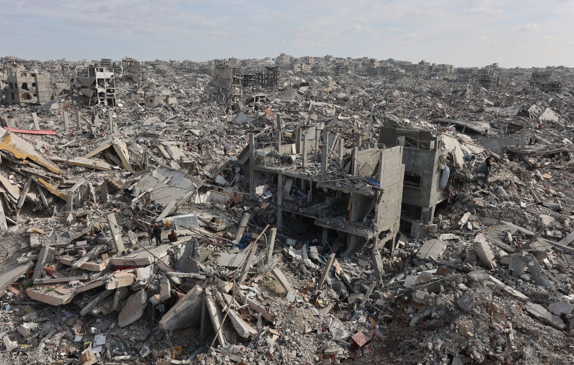 L'image montre un vaste paysage de débris et de bâtiments effondrés, engloutis dans les gravats. On aperçoit des structures en béton brisées et une atmosphère de destruction désolante.