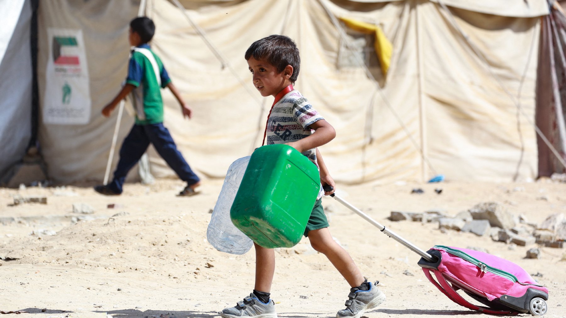 L'image montre un enfant marchant dans un environnement de camp, probablement un camp de réfugiés. Il tire une valise rose avec une poignée, tout en portant un récipient vert dans ses bras. En arrière-plan, on peut voir d'autres enfants et des tentes en toile, symbolisant un cadre de vie précaire. Le sol est poussiéreux et dénudé, ce qui reflète les conditions difficiles de l'endroit. L'enfant a un visage déterminé et semble concentré sur sa tâche.