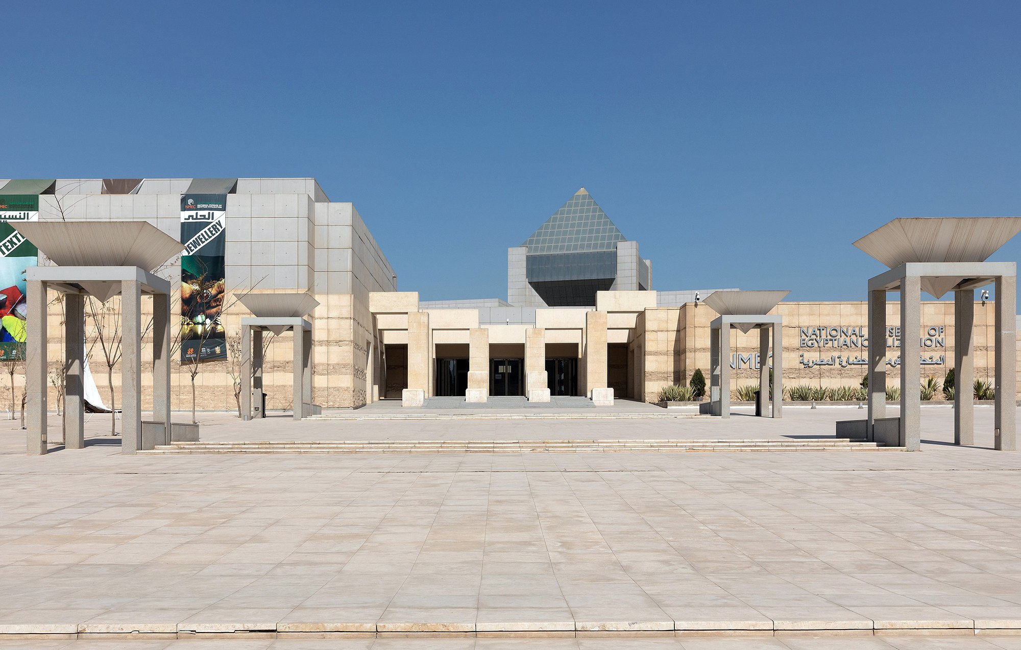 L'image montre l'entrée d'un bâtiment moderne, probablement un musée ou un centre culturel, avec une architecture distinctive. En façade, on voit plusieurs colonnes soutenant un toit en forme de voile, et une grande structure pyramidale se dresse à l'arrière. Les murs sont en pierre beige, donnant un aspect élégant et contemporain. Le ciel est clair, sans nuages, ce qui crée une atmosphère lumineuse et accueillante. Les éléments graphiques sur les murs suggèrent des expositions ou des événements.