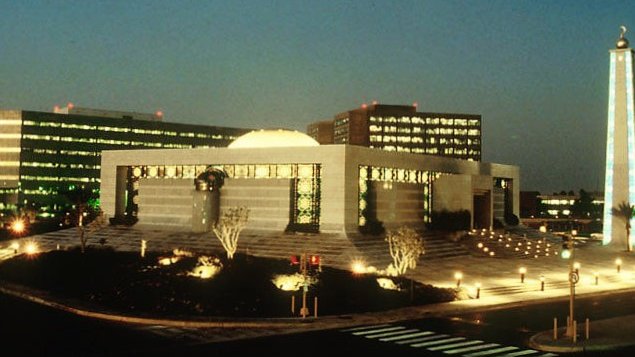 L'image montre un paysage urbain nocturne, avec un bâtiment moderne bien éclairé au centre. Ce bâtiment a une architecture distinctive, avec une coupole et des éléments de design contemporains. À l'arrière-plan, plusieurs autres structures se dressent, leurs fenêtres illuminées créant une ambiance vivante. On peut également voir une grande lune brillante dans le ciel, ajoutant une touche romantique à la scène. Des arbres et des zones paysagées entourent le bâtiment, et des lampadaires éclairent les chemins et les espaces extérieurs, rendant l'ensemble accueillant et esthétique.