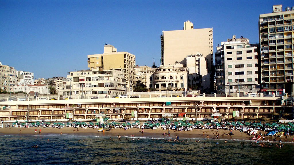 L'image montre une plage animée avec de nombreux baigneurs profitant du soleil et de la mer. On peut voir des parasols disposés sur le sable, ainsi que des hôtels et des immeubles en arrière-plan. L'architecture des bâtiments est variée, avec des structures anciennes côtoyant des constructions plus modernes. Le ciel est dégagé, et l'atmosphère est vivante, typique d'une destination balnéaire populaire.