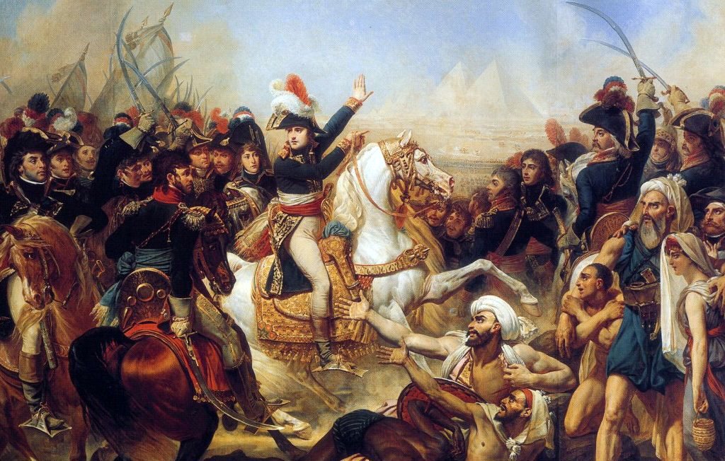 L'image représente une scène historique chargée d'émotions, probablement liée à la campagne d'Égypte de Napoléon Bonaparte. Au premier plan, Napoléon est sur un cheval blanc, vêtu de son uniforme militaire, semblant diriger ses troupes. On aperçoit des soldats français en uniforme, ainsi que des combattants locaux, ce qui indique un affrontement. À l'arrière-plan, les pyramides d'Égypte se dressent majestueusement, ajoutant une dimension emblématique à la scène. L'atmosphère est intense, reflétant le chaos et la violence de la bataille.