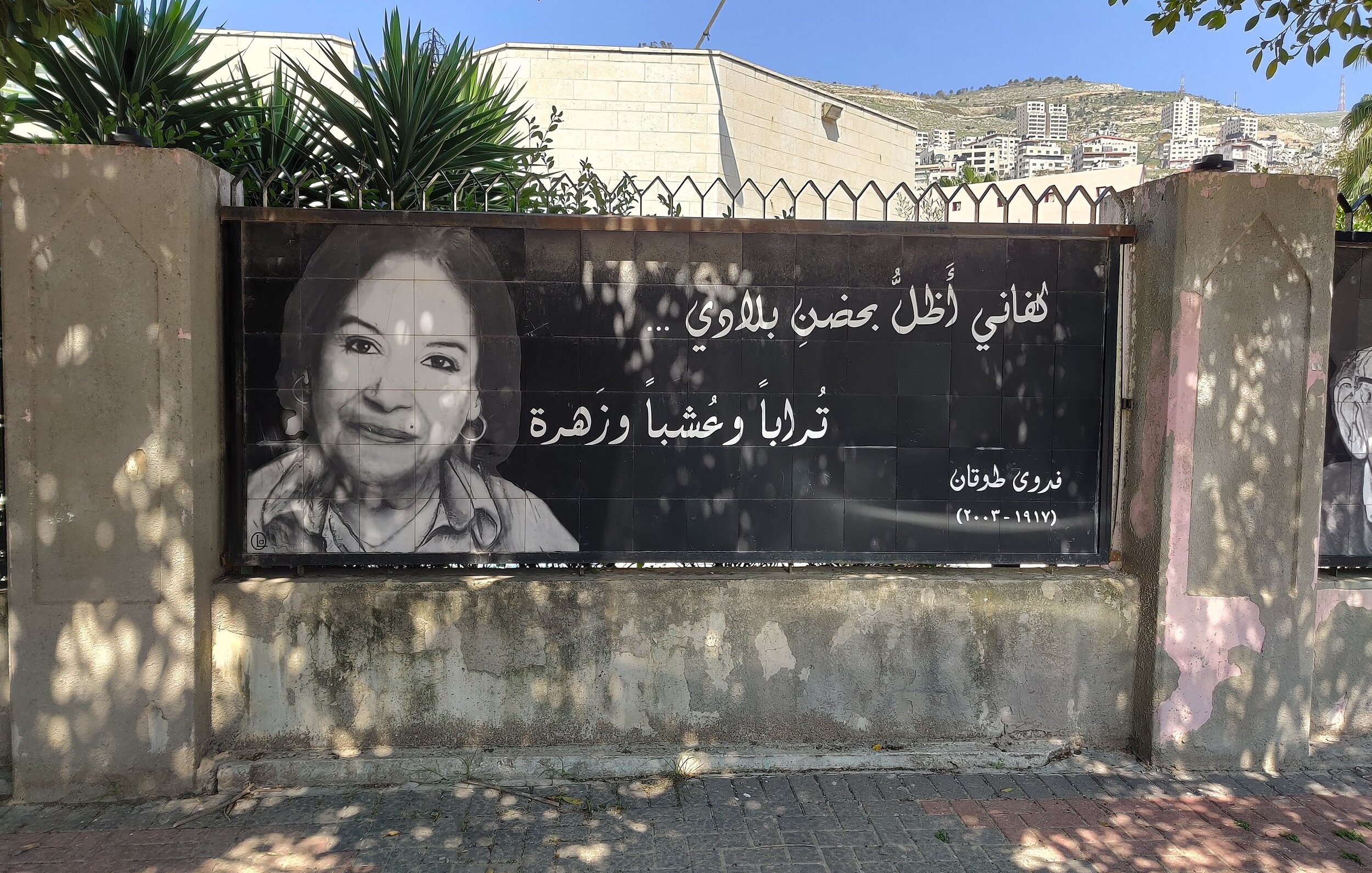 L'image montre une grande affiche accrochée à une clôture. Elle présente un portrait en noir et blanc d'une femme souriante. À côté de son visage, on trouve une citation en arabe, ainsi qu'un nom et une date en bas. L'arrière-plan montre des bâtiments et des végétaux, indiquant que l'affiche est placée dans un environnement urbain. L'ensemble véhicule une ambiance d'hommage ou de souvenir.