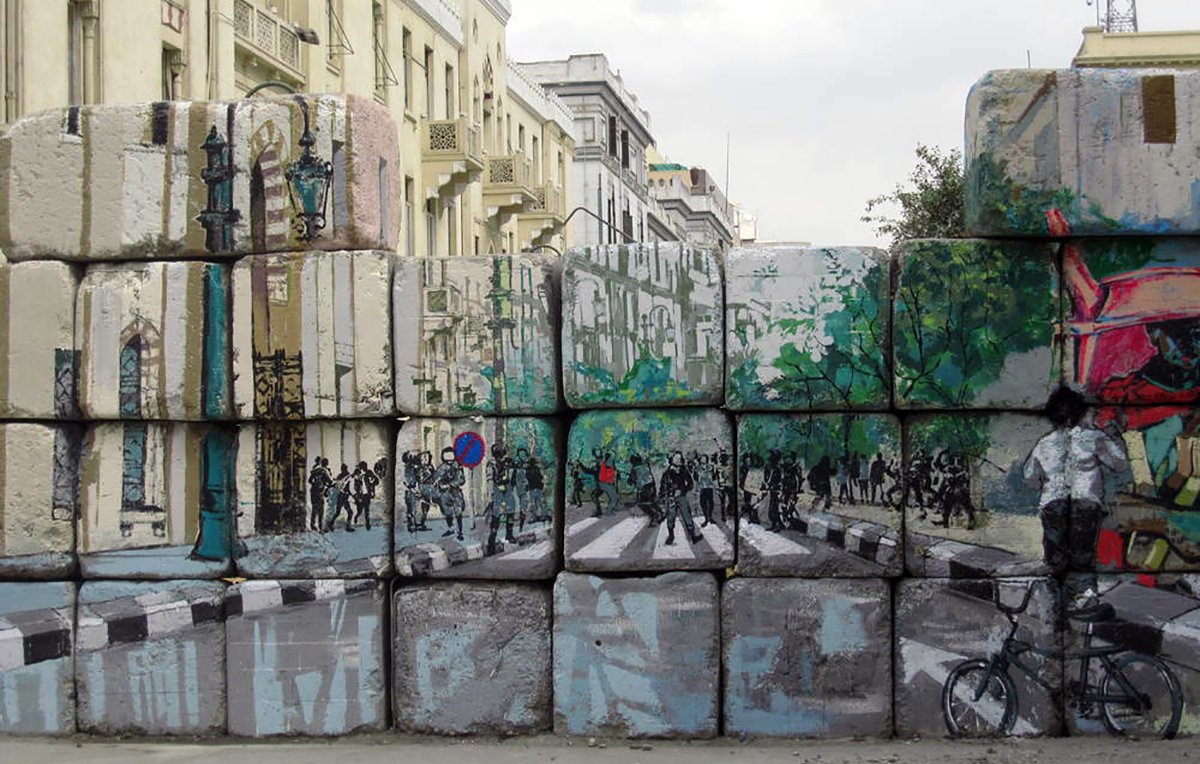 L'image montre un mur de blocs de béton, sur lequel sont peints des paysages urbains. La peinture représente une rue avec des arbres, des piétons et des cyclistes. On peut voir des silhouettes de personnes et des éléments de la ville, comme des lampadaires. Ce contraste entre le mur brut et l'art coloré apporte une touche dynamique à l'environnement urbain. Le ciel est nuageux, ajoutant une atmosphère un peu dramatique à la scène.