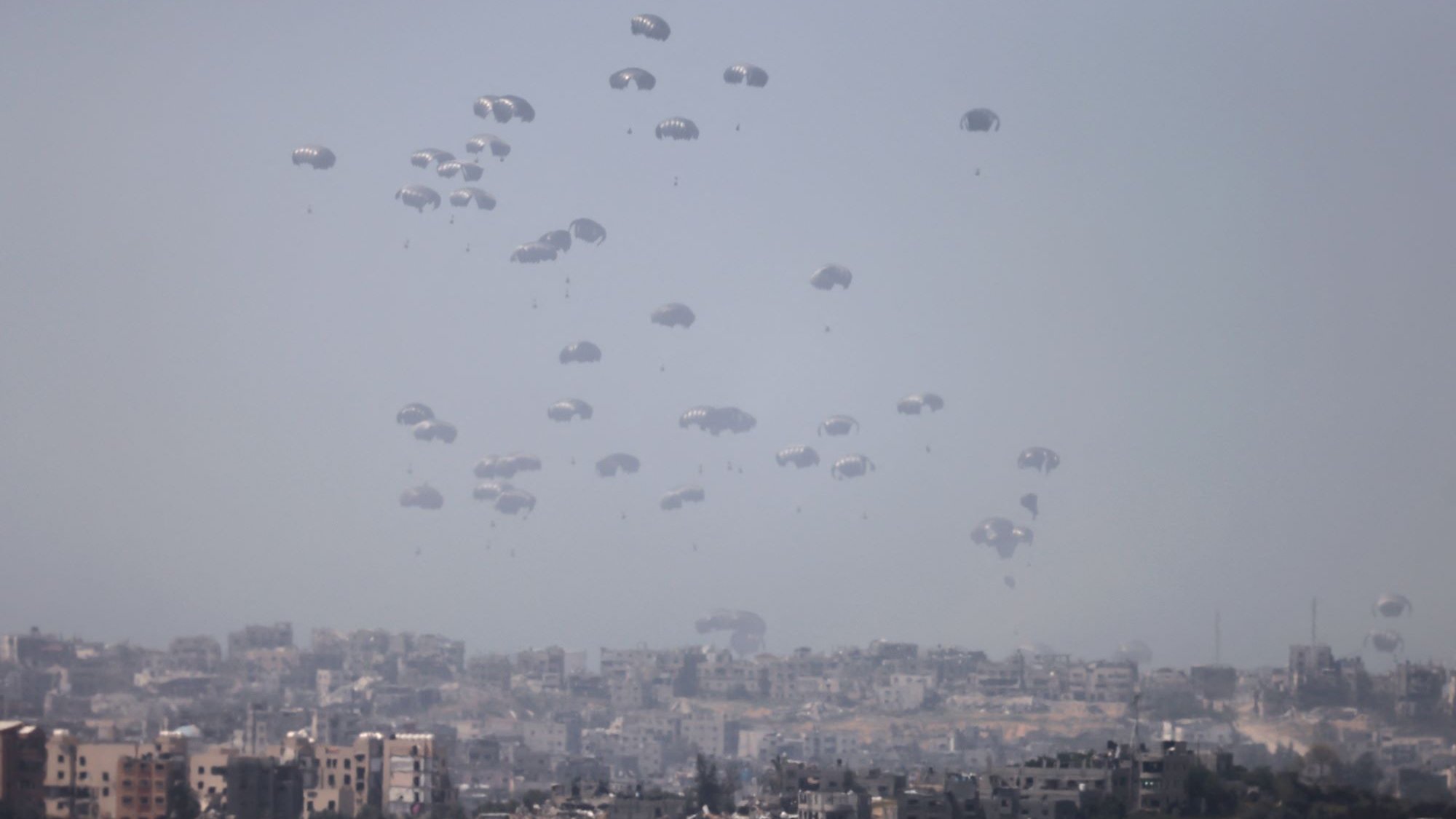 L'image montre un paysage urbain où de nombreux parachutes descendent lentement vers le sol. On peut voir une ville en arrière-plan, avec des bâtiments disposés de manière variable. L'atmosphère semble brumeuse, ce qui donne un aspect à la fois dramatique et intrigant à la scène. Les parachutes indiquent probablement une opération militaire ou humanitaire, créant un contraste avec l'environnement urbain.