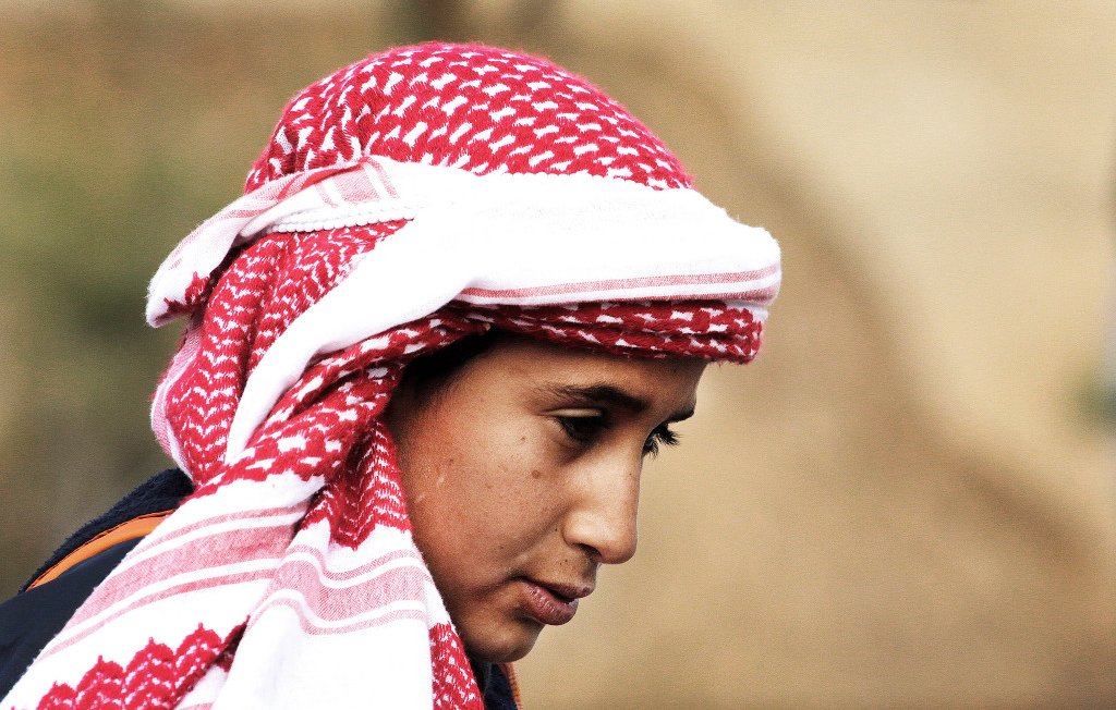 L'image montre un jeune homme portant un keffieh, un traditionnel foulard arabe, qui est souvent orné de motifs rouges et blancs. Son expression semble pensive, et il a les cheveux bruns. L'arrière-plan est flou, indiquant un environnement naturel ou rural. Son regard est tourné légèrement vers le bas, ce qui ajoute une certaine introspection à son apparence.