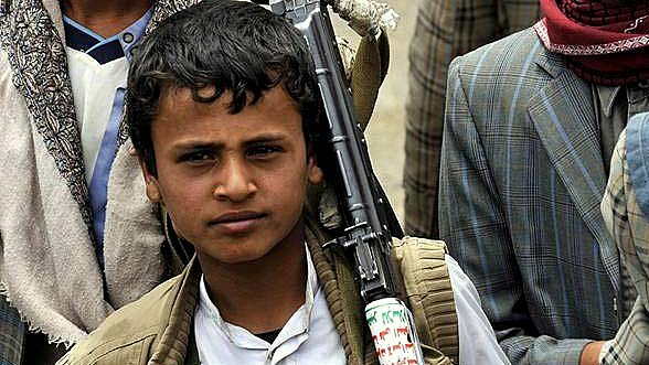 L'image montre un jeune garçon, probablement dans un contexte difficile. Il porte des vêtements simples et a un fusil en bandoulière. Son regard est sérieux, et il semble faire partie d'un groupe plus large, qui est à l'arrière-plan. Les vêtements des autres personnes indiquent un environnement rural ou de conflit. L'atmosphère générale de l'image témoigne d'un contexte de tension.