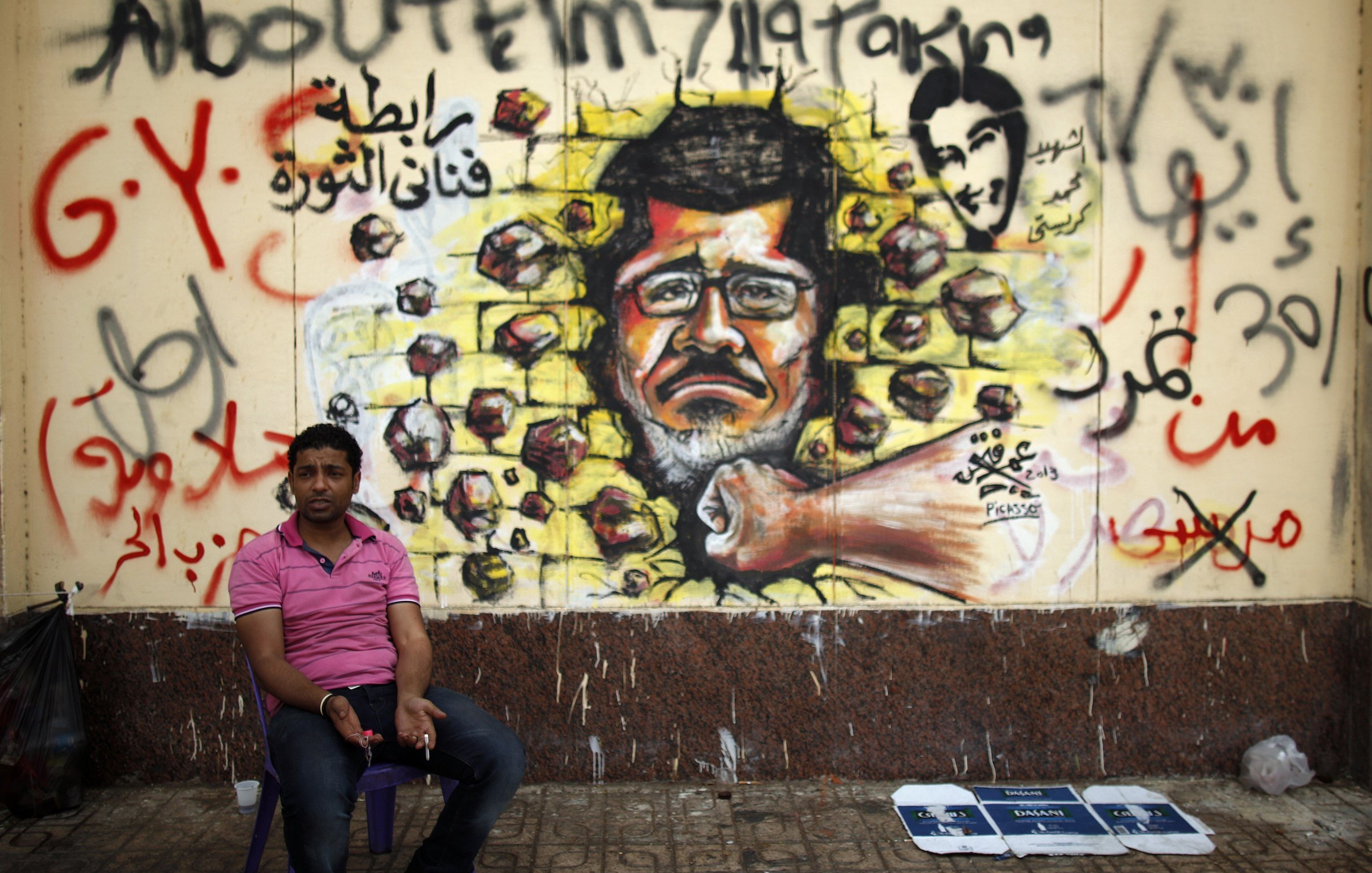 L'image montre un homme assis sur une petite chaise, devant un mur qui est recouvert d'une fresque murale. Cette fresco représente un portrait d'un homme au visage sérieux, avec des roches projetées autour de lui, symbolisant peut-être la résistance ou la lutte. Le mur est également couvert d'inscriptions en arabe et peut-être en d'autres langues, ajoutant un contexte politique ou social à la scène. L'environnement semble urbain, avec un sol en pavés et des déchets visibles à proximité.