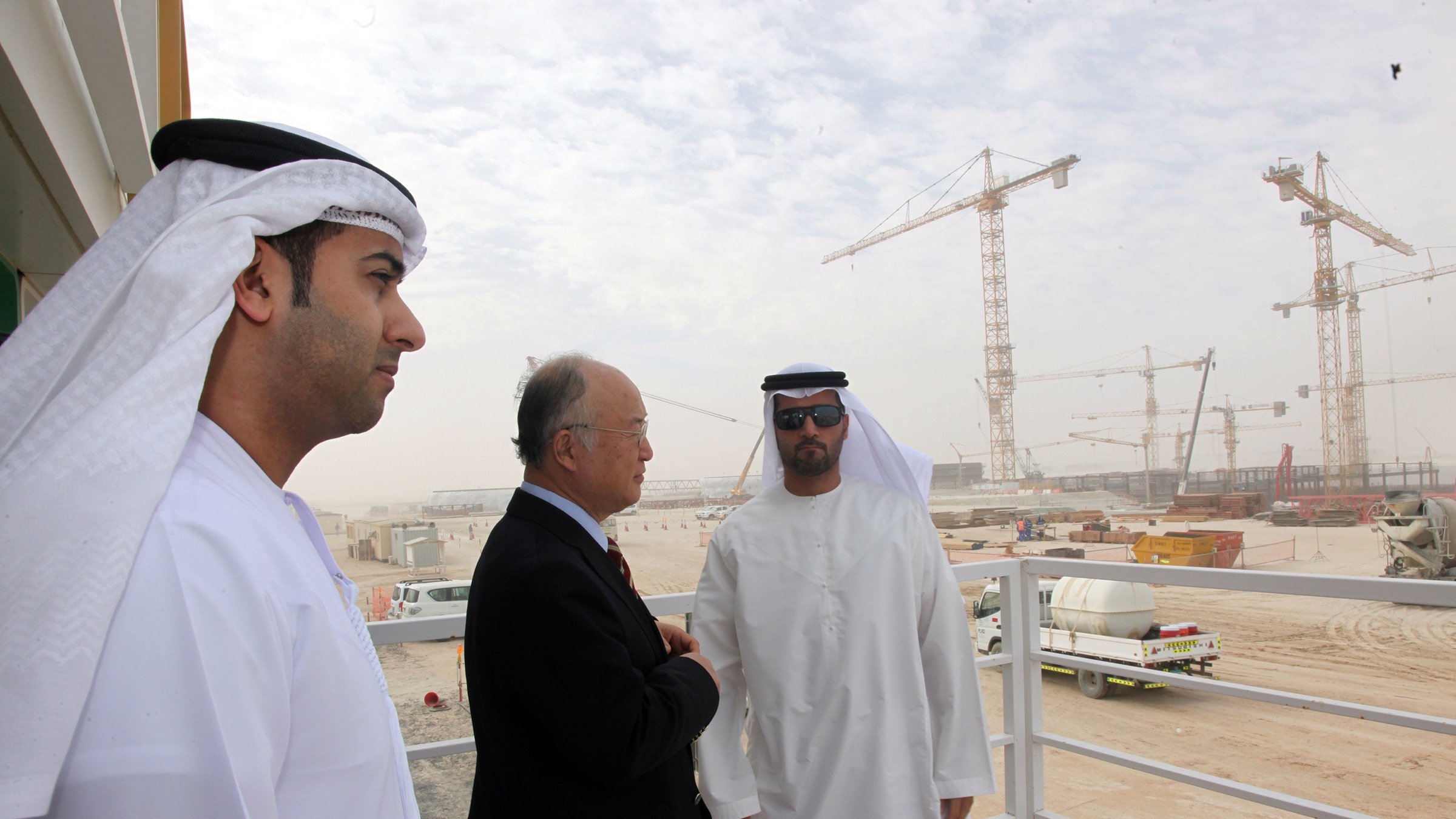 L'image montre trois hommes, apparemment en discussion, sur un balcon ou une plateforme. Ils sont vêtus de vêtements traditionnels des Émirats, comprenant des kandouras et des ghutras. En arrière-plan, on peut voir un site de construction avec des grues et des véhicules, suggérant un projet de développement en cours. L'environnement est poussiéreux et légèrement brumeux, typique des zones de construction dans des climats chauds. Les hommes semblent sérieux et engagés dans leur conversation.