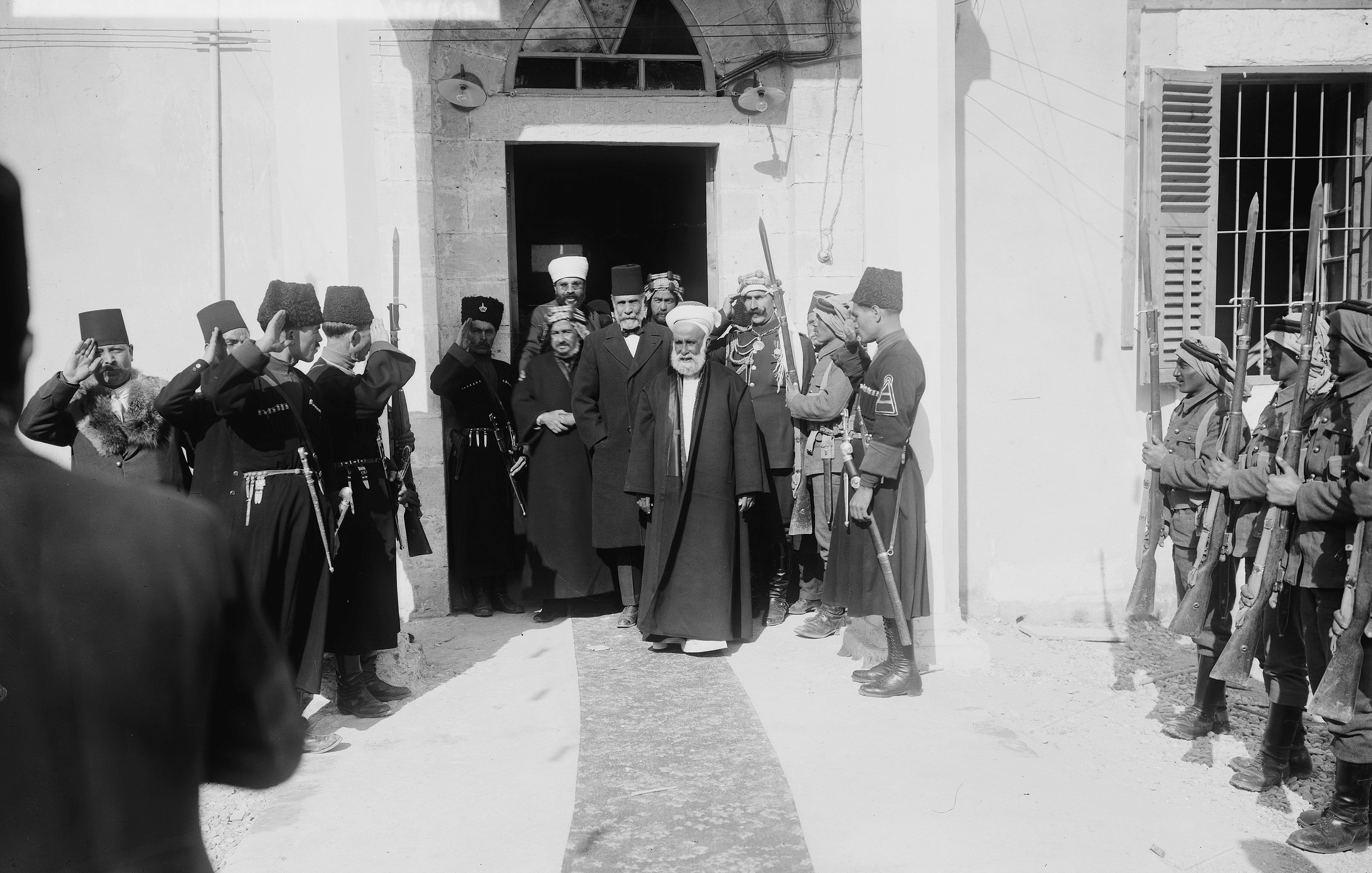 L'image montre un groupe de personnes en costume traditionnel, saluant respectueusement un homme au centre qui semble être une figure de proue. L'ambiance est formelle et cérémonieuse. On peut voir des soldats en uniforme tenant des armes et des dignitaires en robes longues. L'architecture en arrière-plan indique que la scène se déroule probablement dans un bâtiment important ou historique. Les expressions des personnes présentent témoignent d'un moment solennel ou d'une cérémonie officielle.