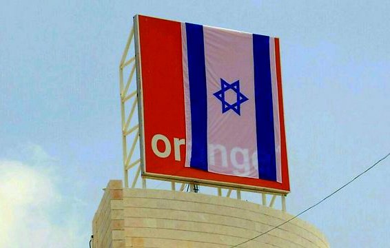 L'image montre un panneau affichant le logo de la marque "Orange", sur lequel est drapé un grand drapeau israélien. Le fond semble être un ciel clair, et le drapeau présente des bandes horizontales en bleu et blanc avec l'étoile de David au centre. Le tout donne une impression d'affichage public dans un environnement urbain.