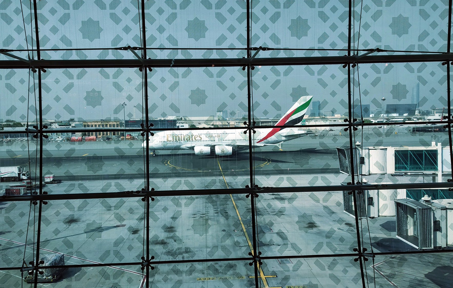 L'image montre l'extérieur d'un aéroport, avec un avion de la compagnie Emirates stationné sur le tarmac. La vue est prise à travers des grandes fenêtres en verre décorées d'un motif géométrique. On peut apercevoir d'autres avions, des bagages et des installations aéroportuaires dans le fond. L'atmosphère semble ensoleillée et on ressent un environnement moderne et animé.