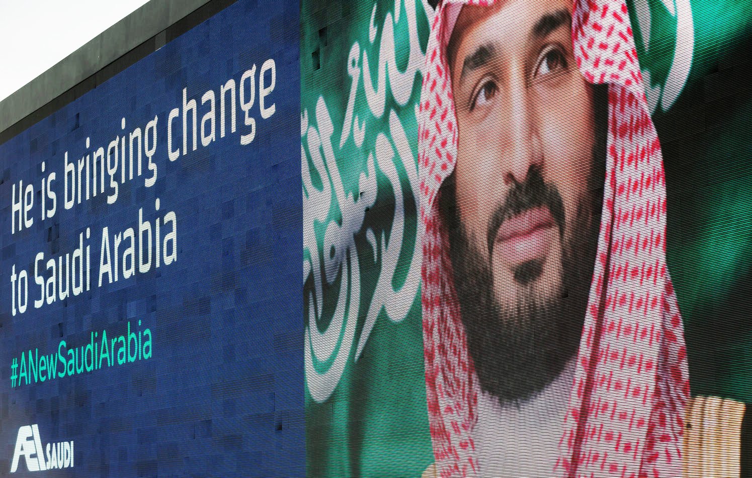 L'image montre une grande affiche murale présentant un homme en tenue traditionnelle saoudienne, avec un keffieh sur la tête. À côté de son visage, il y a un message en anglais qui dit "He is bringing change to Saudi Arabia" et un hashtag "#NewSaudiArabia". L'arrière-plan de l'affiche est bleu et le texte blanc. Cela évoque un thème de changement et de modernisation en Arabie Saoudite.