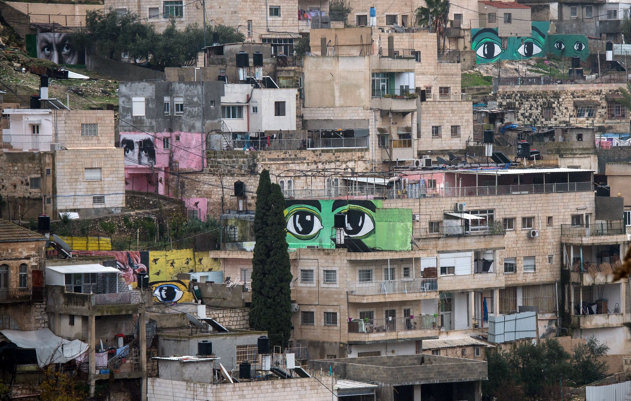 L'image montre un paysage urbain d'une ville, où plusieurs bâtiments sont alignés sur une colline. À travers ces constructions, on peut voir de grandes peintures murales représentant des yeux, aux couleurs vives, qui ajoutent une dimension artistique à cet environnement. Les bâtiments varient en couleurs et en textures, allant du beige au rose, et certains semblent en cours de rénovation. L'atmosphère générale évoque un mélange de quotidien et de créativité, avec une forte présence d'art urbain.
