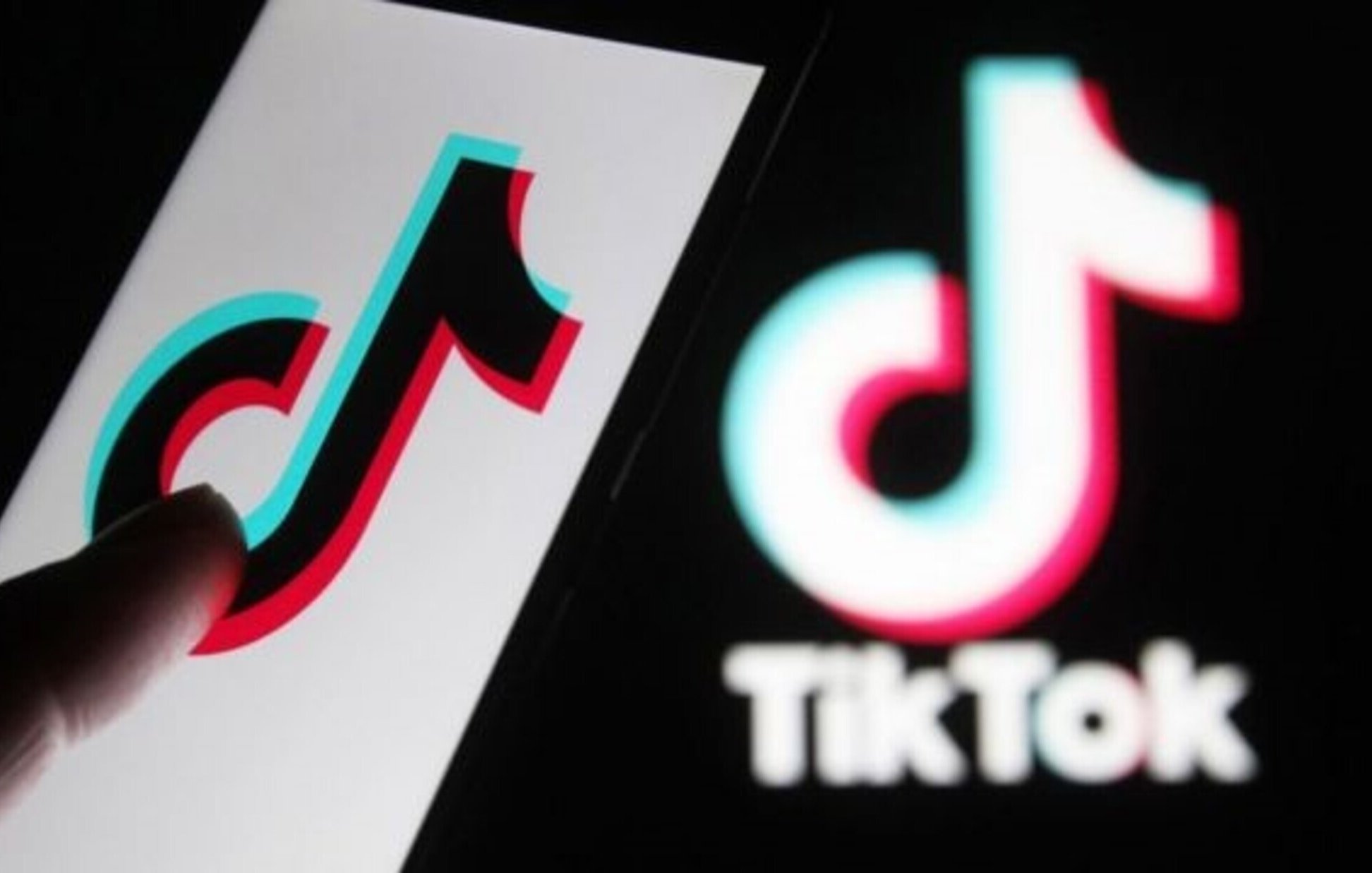 L'image montre un smartphone avec le logo de TikTok affiché à l'écran. On peut voir un doigt en train de toucher ou interagir avec le logo. À l'arrière-plan, il y a le logo de TikTok dans un style similaire, mais peut-être moins détaillé. Les couleurs principales du logo sont le noir, le rouge et le bleu, créant un effet visuel attrayant.