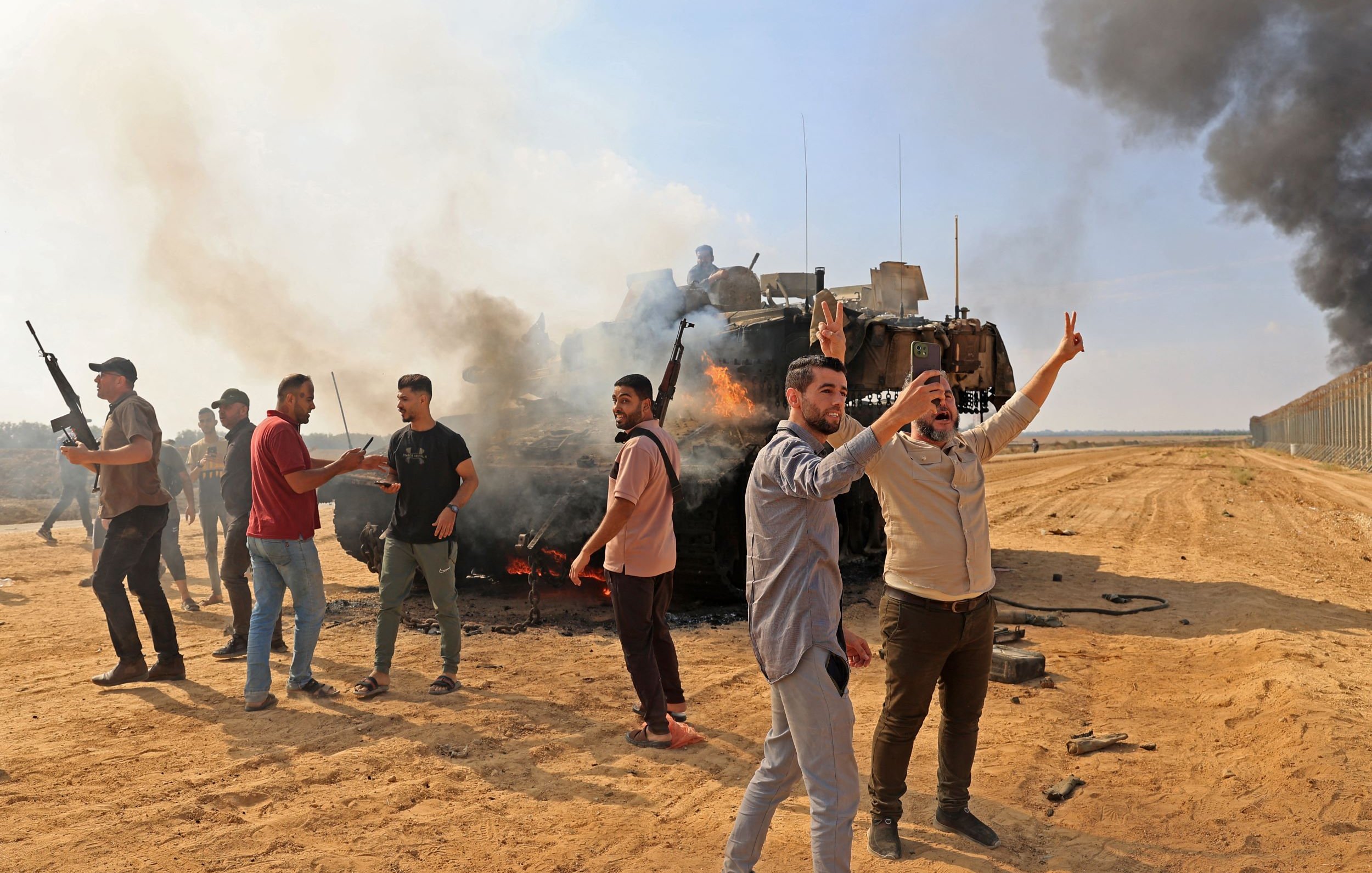 L'image montre un groupe de personnes dans un environnement de conflit. Au premier plan, plusieurs hommes expriment des gestes de victoire ou de protestation, tandis que certains portent des armes. En arrière-plan, on peut voir un véhicule militaire en feu, entouré de fumée noire. Le sol est sablonneux, et le paysage général évoque une situation de tension ou de guerre. L'ambiance est tumultueuse, reflétant un moment de confrontation ou de manifestation.